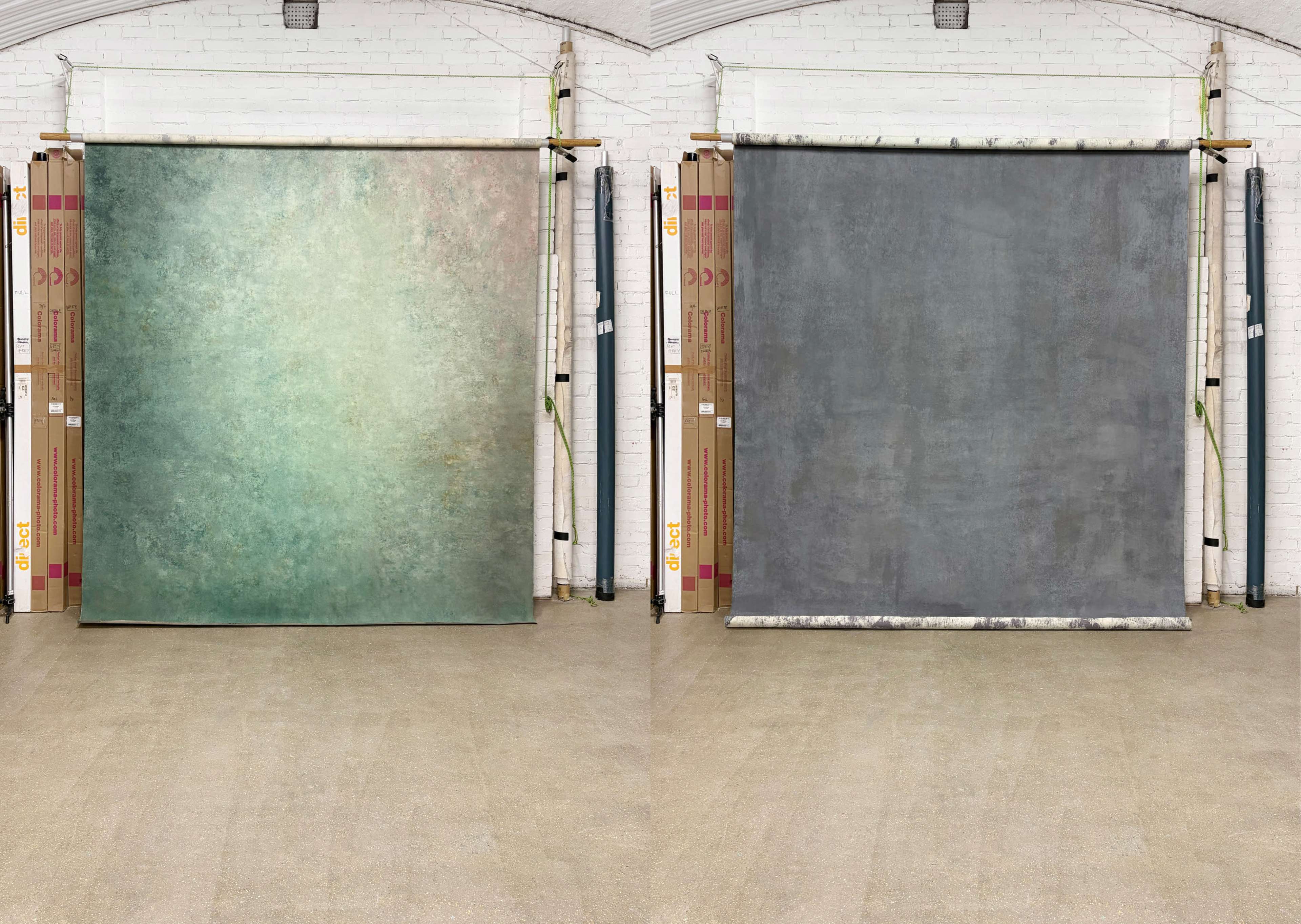 The image shows two large backdrops displayed in a studio, one with a textured green surface and the other with a solid gray surface, both set against a neutral floor and white brick wall.