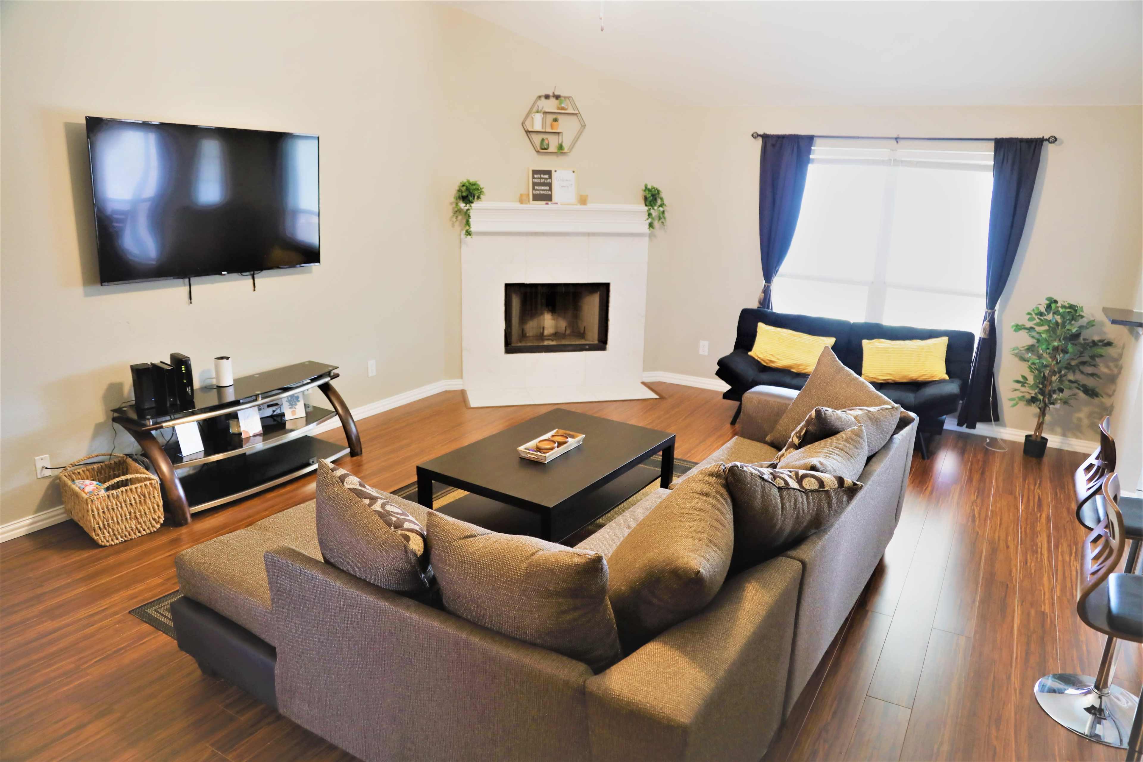 The living room features a gray sectional sofa, a black coffee table, a television mounted on the wall, and a fireplace surrounded by light-colored walls and wooden flooring.