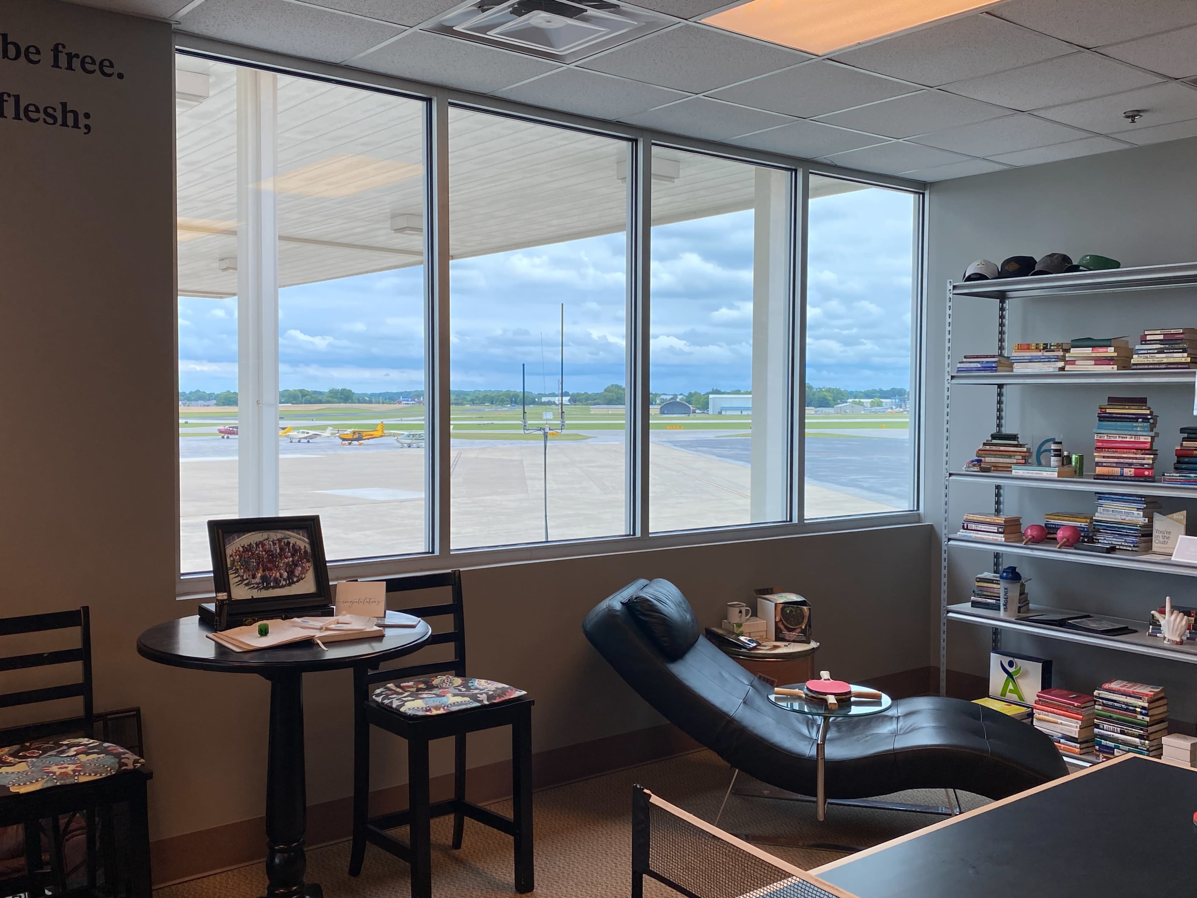 A lounge area with a chaise lounge and shelves of books overlooks an airport runway through large windows.