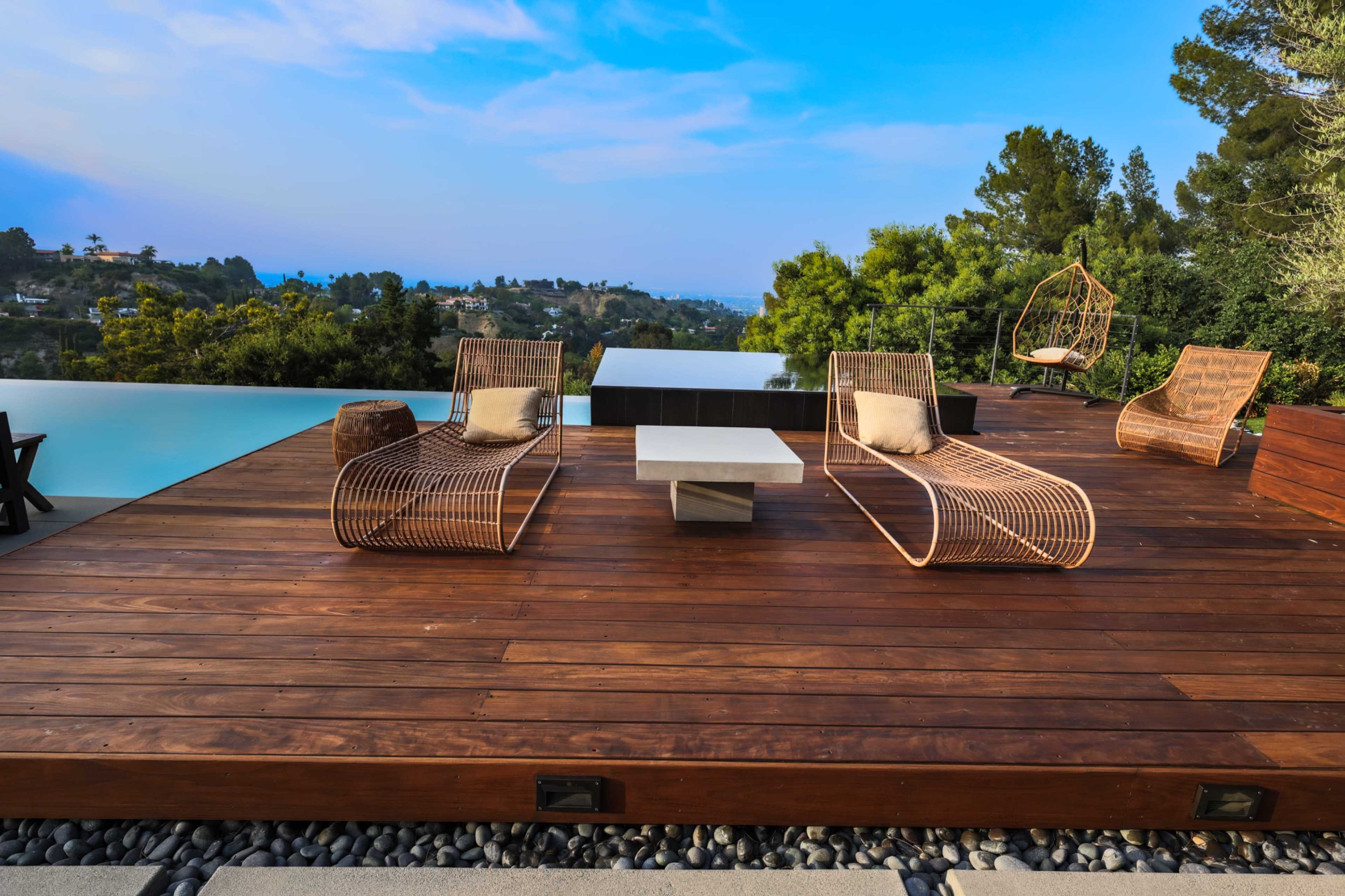 A modern outdoor patio features two curved lounge chairs, a small table, and a hanging chair, all set on a wooden deck overlooking a scenic landscape.