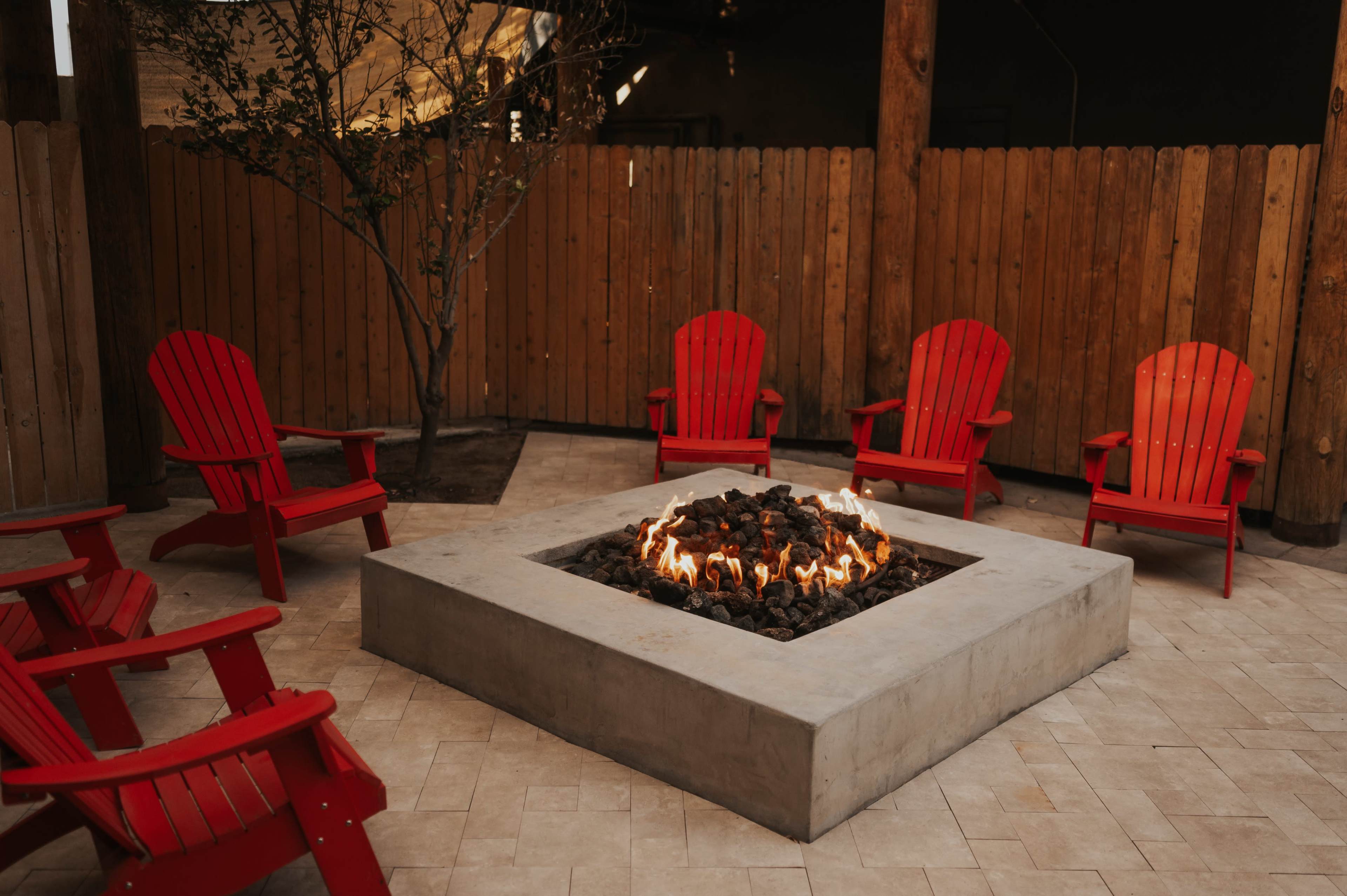 The image shows a circular concrete fire pit surrounded by five red Adirondack chairs set in a fenced outdoor space.
