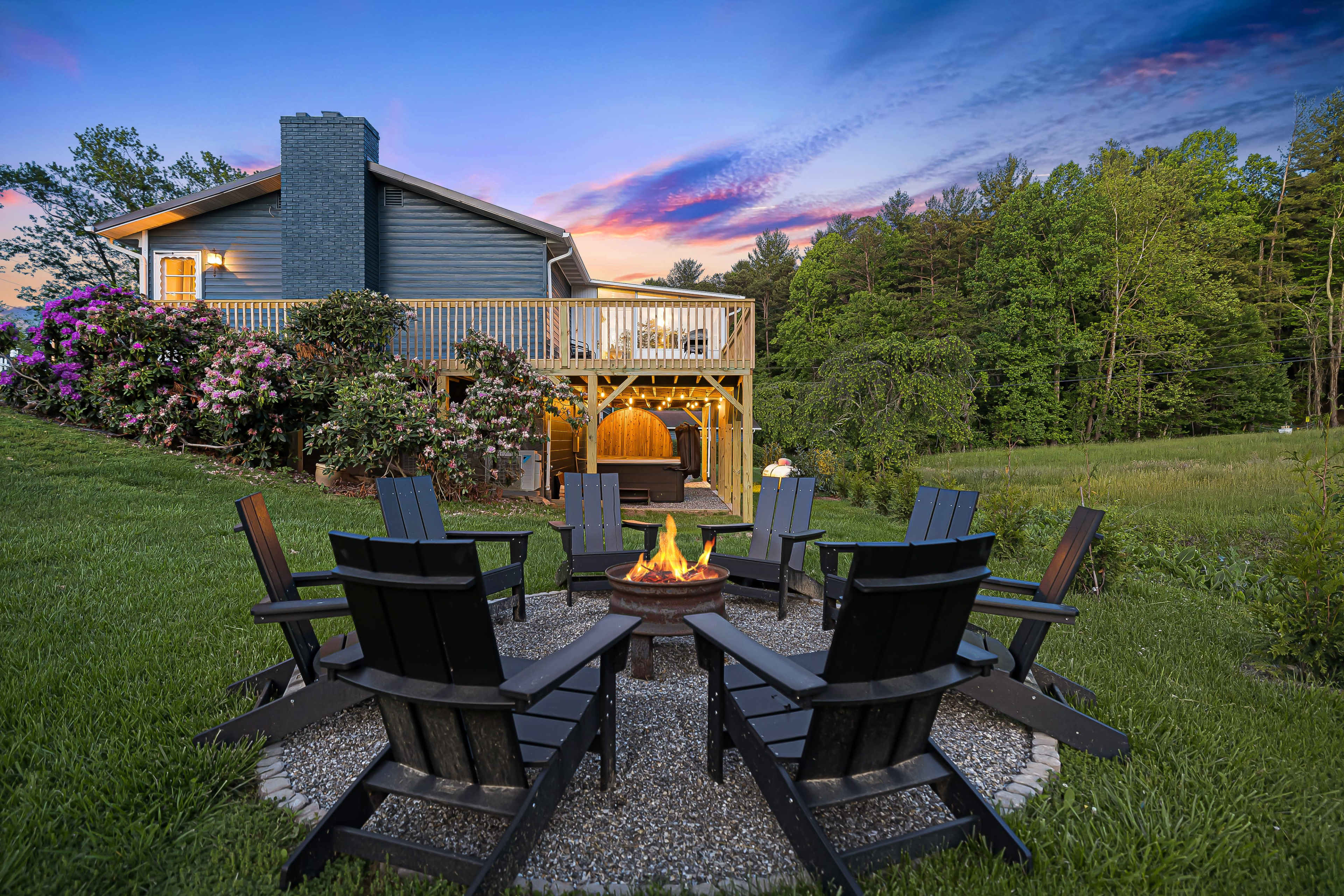 A circular arrangement of black Adirondack chairs surrounds a fire pit in a backyard with a house and a deck in the background, illuminated by a sunset sky.