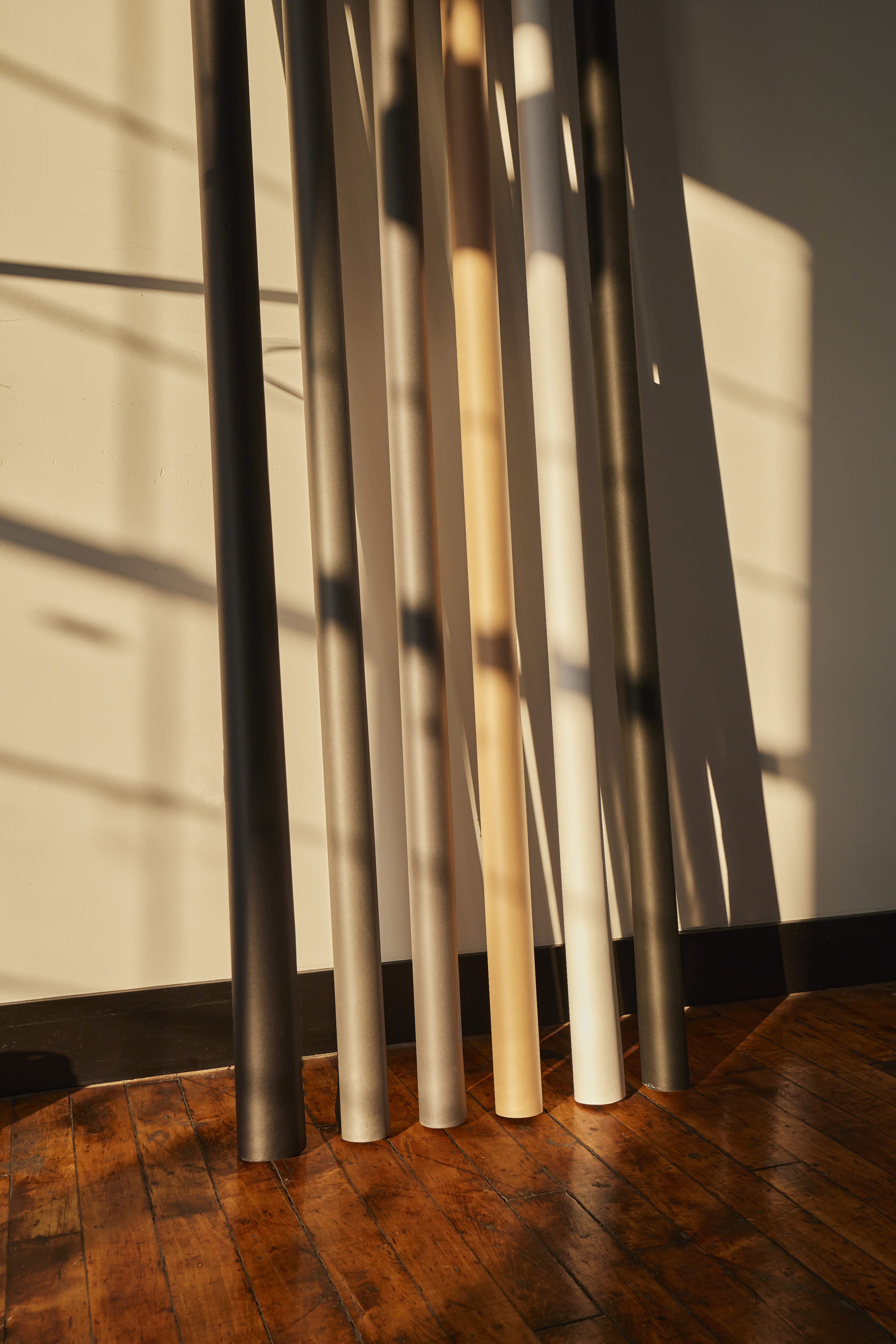 Several vertical rolls of paper standing on a wooden floor, casting shadows on the wall as sunlight streams through a nearby window.