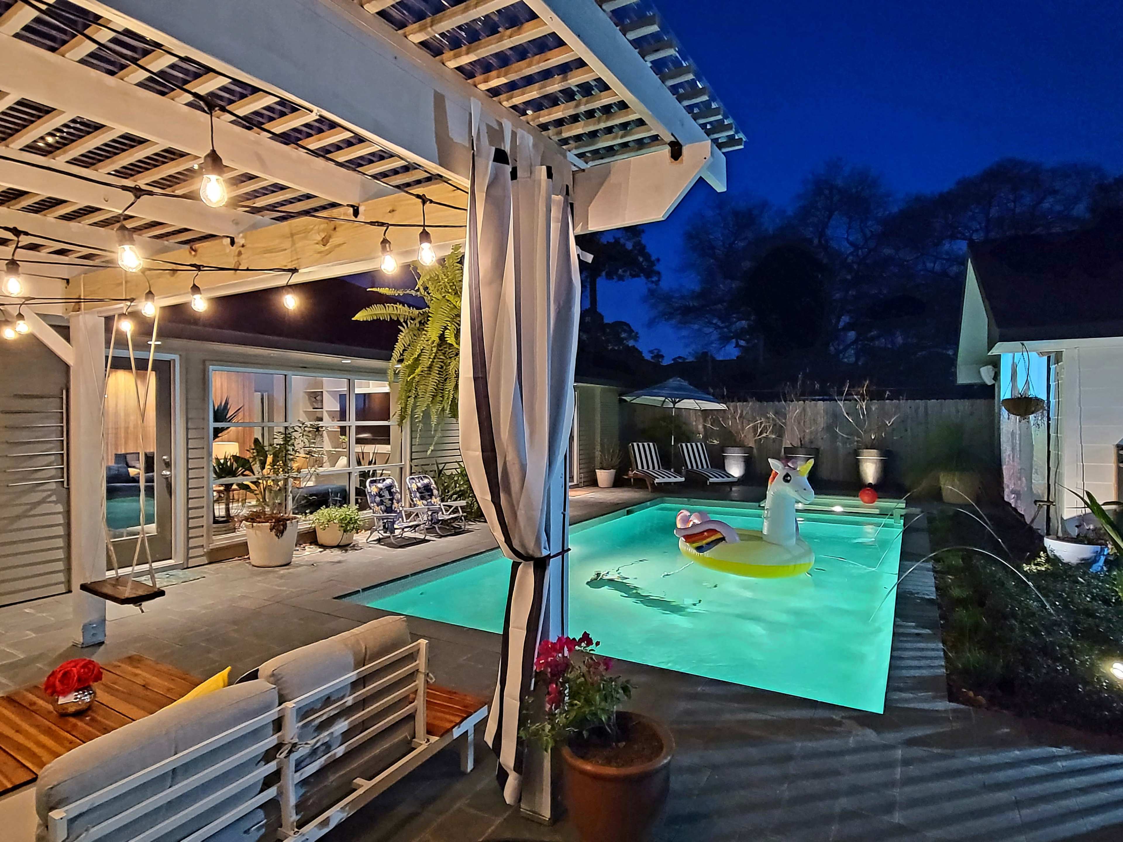 The scene depicts a well-lit backyard pool area at night, featuring a turquoise pool with a large inflatable unicorn and surrounded by comfortable seating and decorative plants.