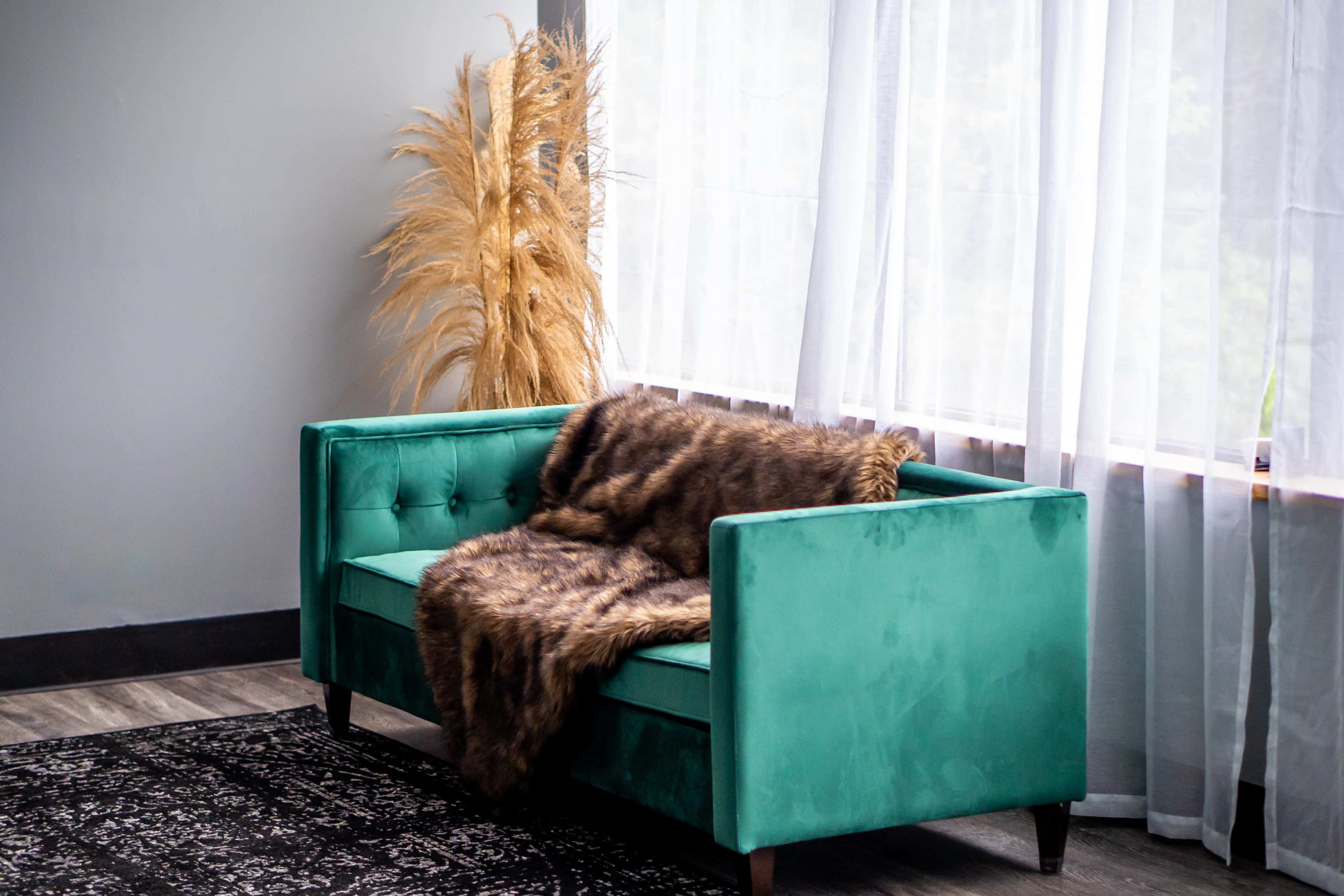 A teal velvet sofa with a fur throw rests against a large window covered by sheer curtains, alongside a decorative arrangement of tall dried plants.