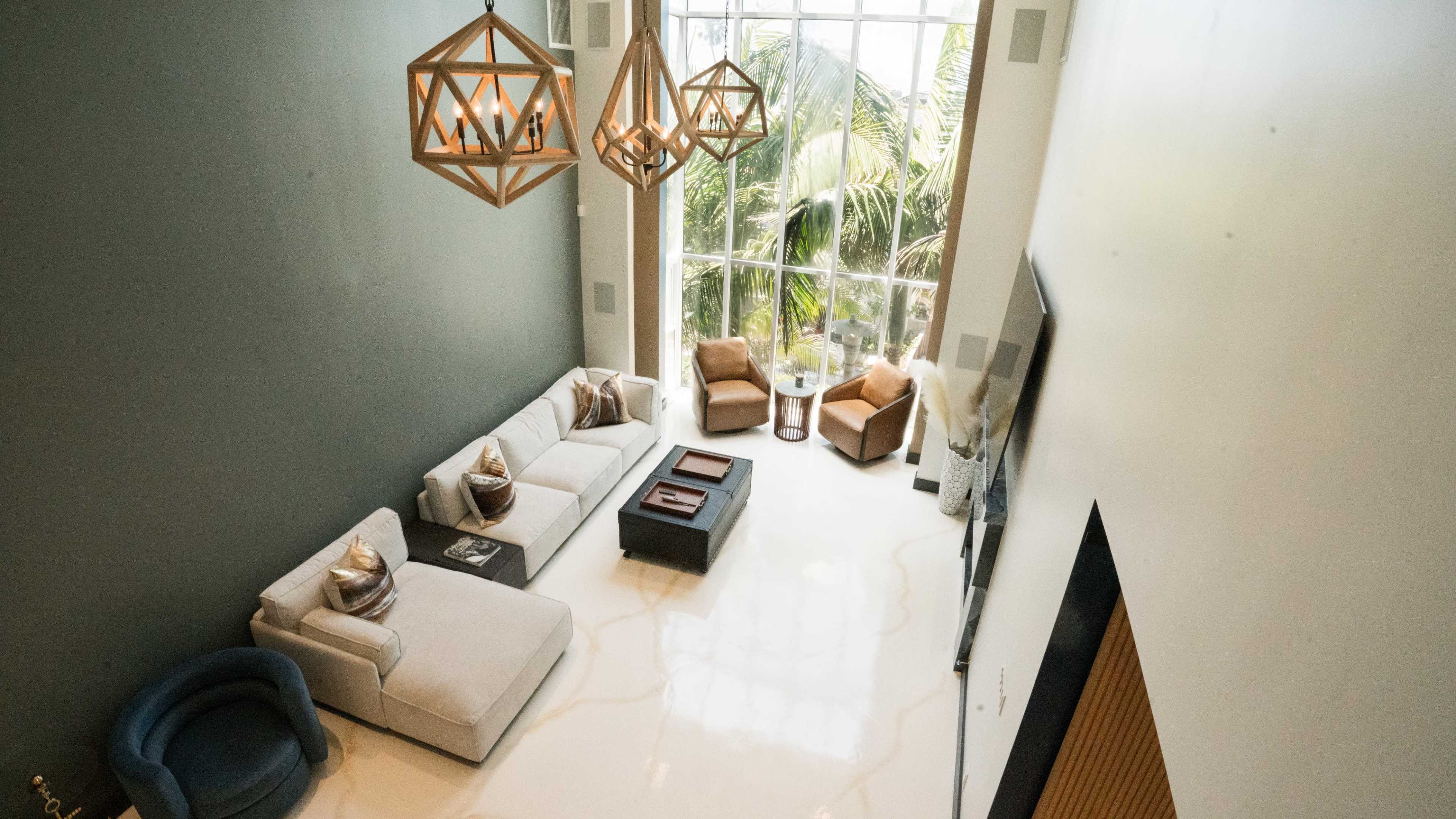 The image shows a spacious living area with large windows, a modern sofa, two armchairs, and decorative light fixtures.