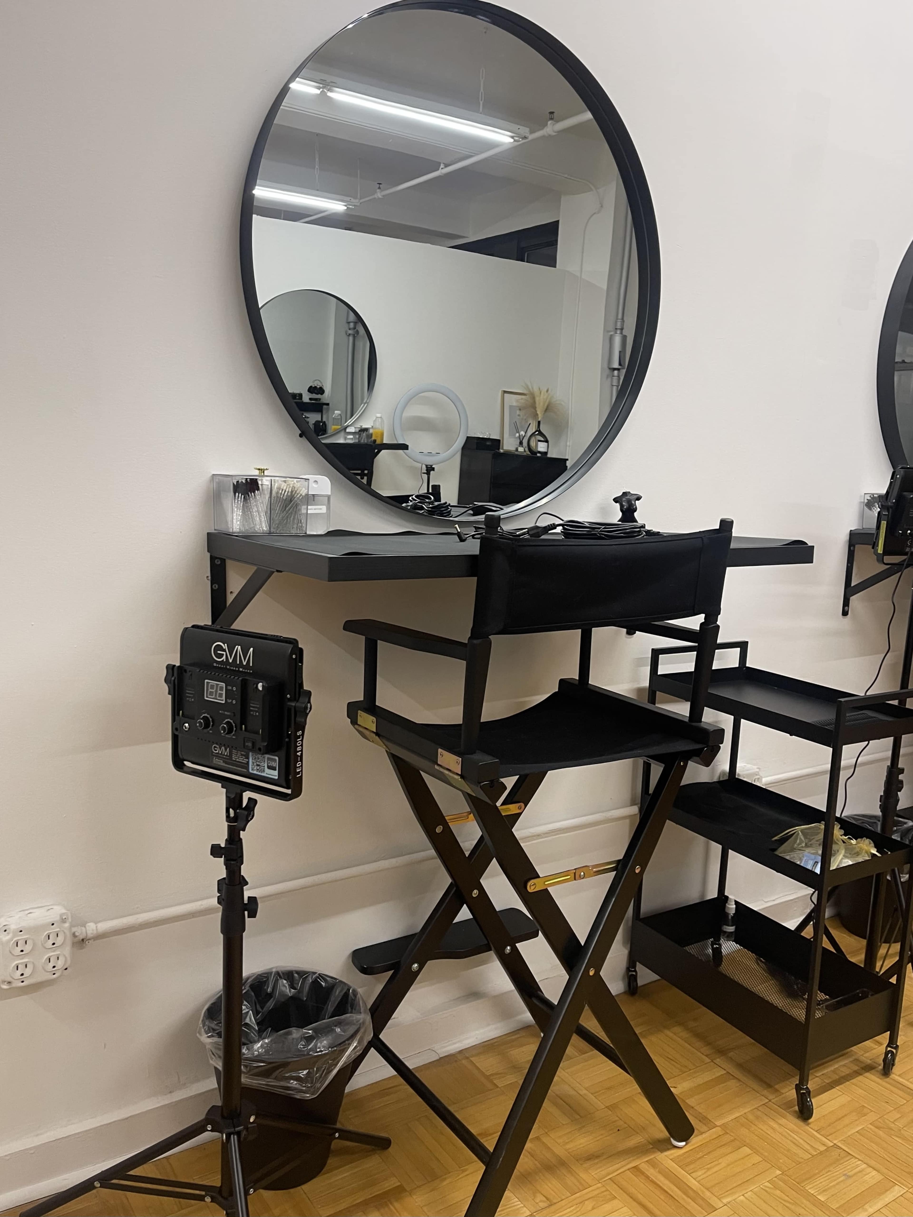 A makeup station featuring a round mirror, a black folding chair, and a small cart, set against a plain white wall.