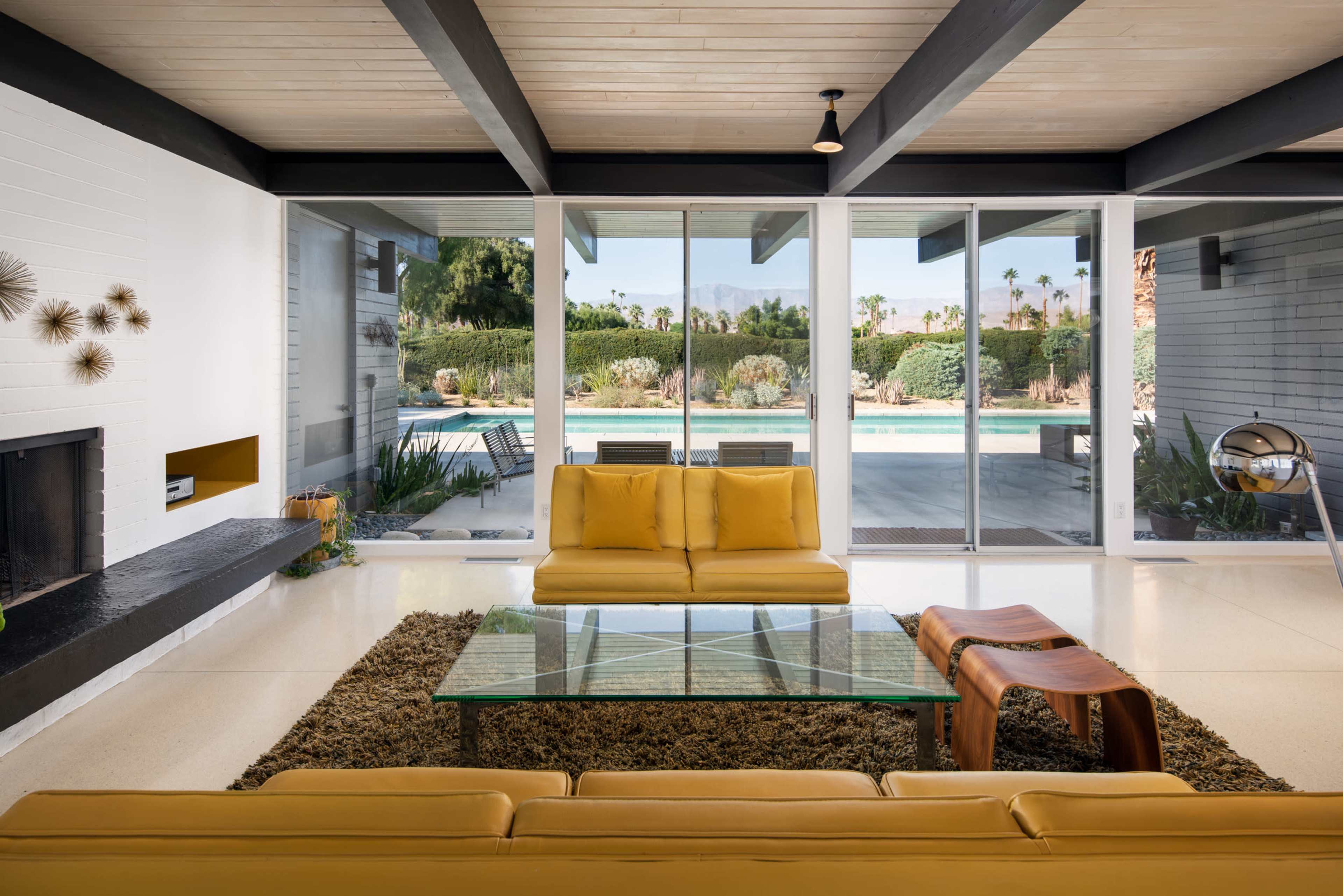 A modern living room featuring a yellow sofa, a glass coffee table, and large windows overlooking a pool area with landscaped greenery.