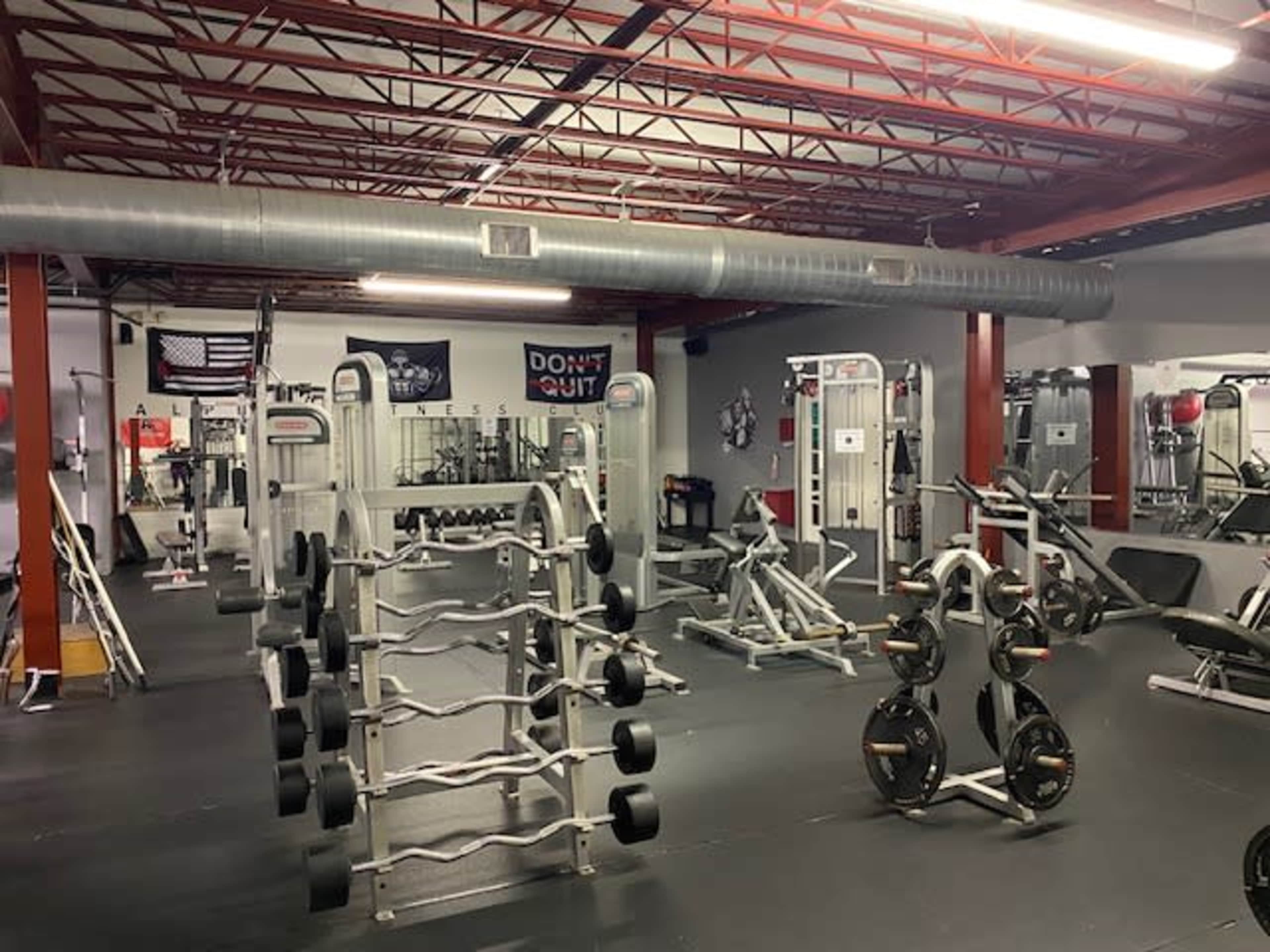 A well-equipped gym filled with various workout machines and free weights, with red and gray walls and a visible American flag in the background.