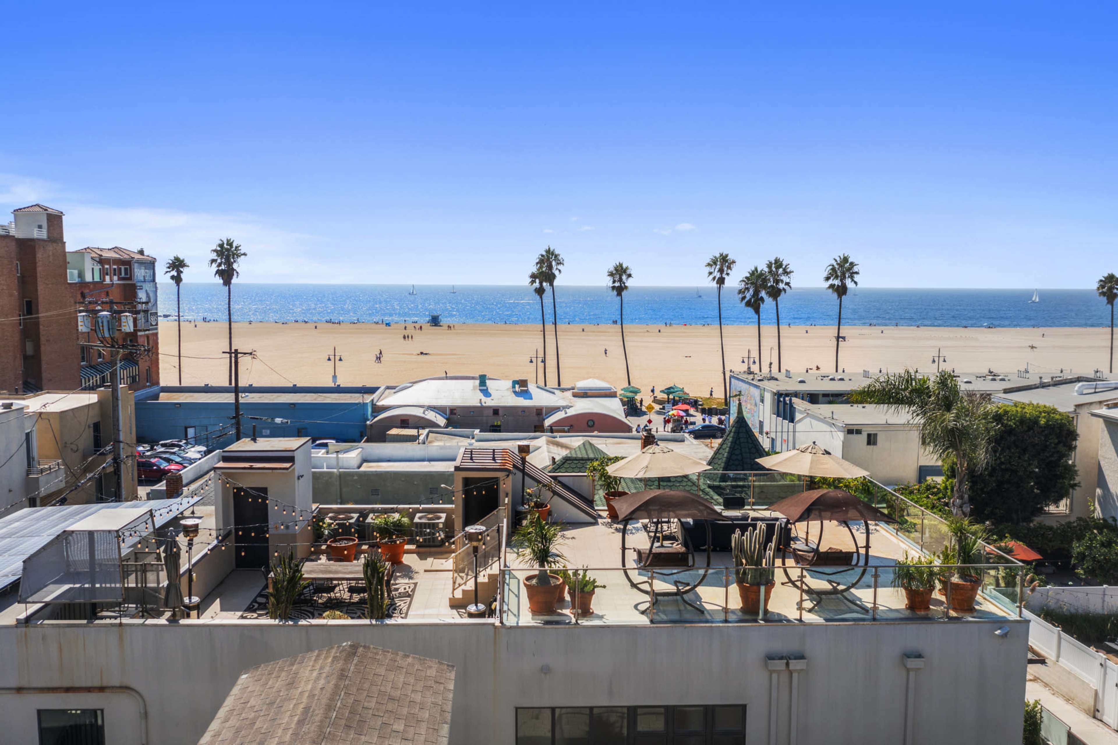 A rooftop view overlooking a beach with palm trees, beachgoers, and colorful structures on the sand.