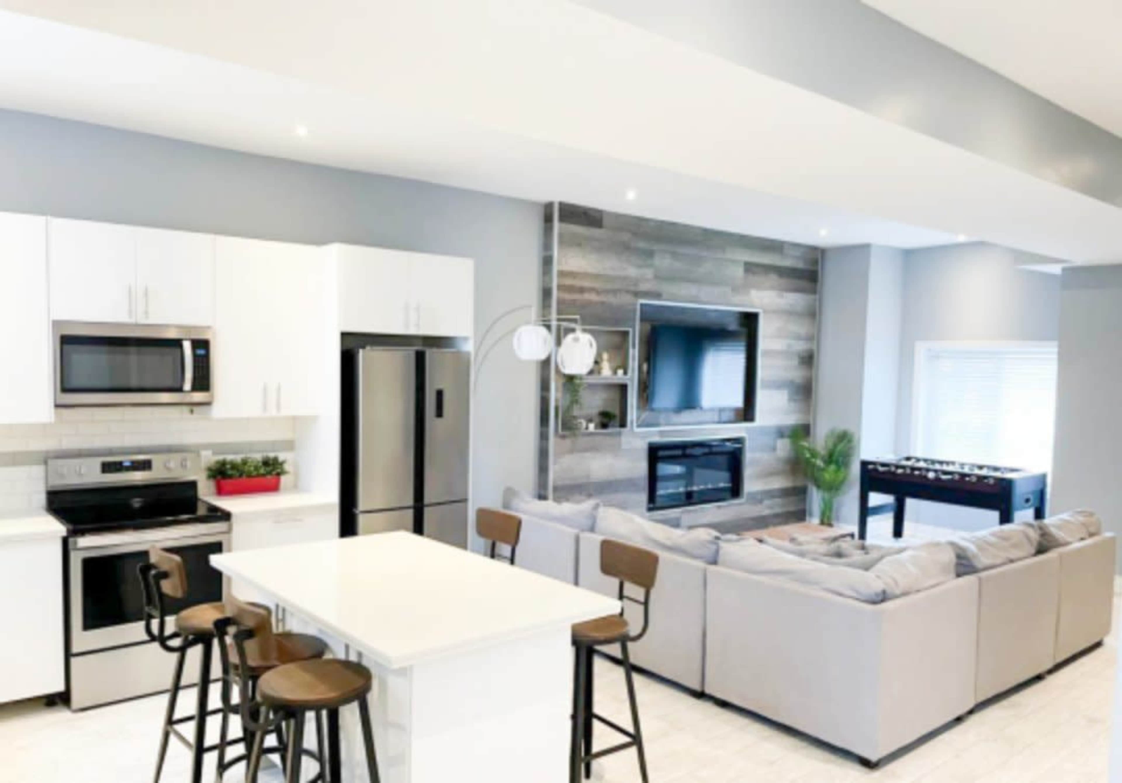 The image shows a modern kitchen and living area featuring white cabinets, stainless steel appliances, a large sectional couch, and a wall-mounted television above a fireplace.