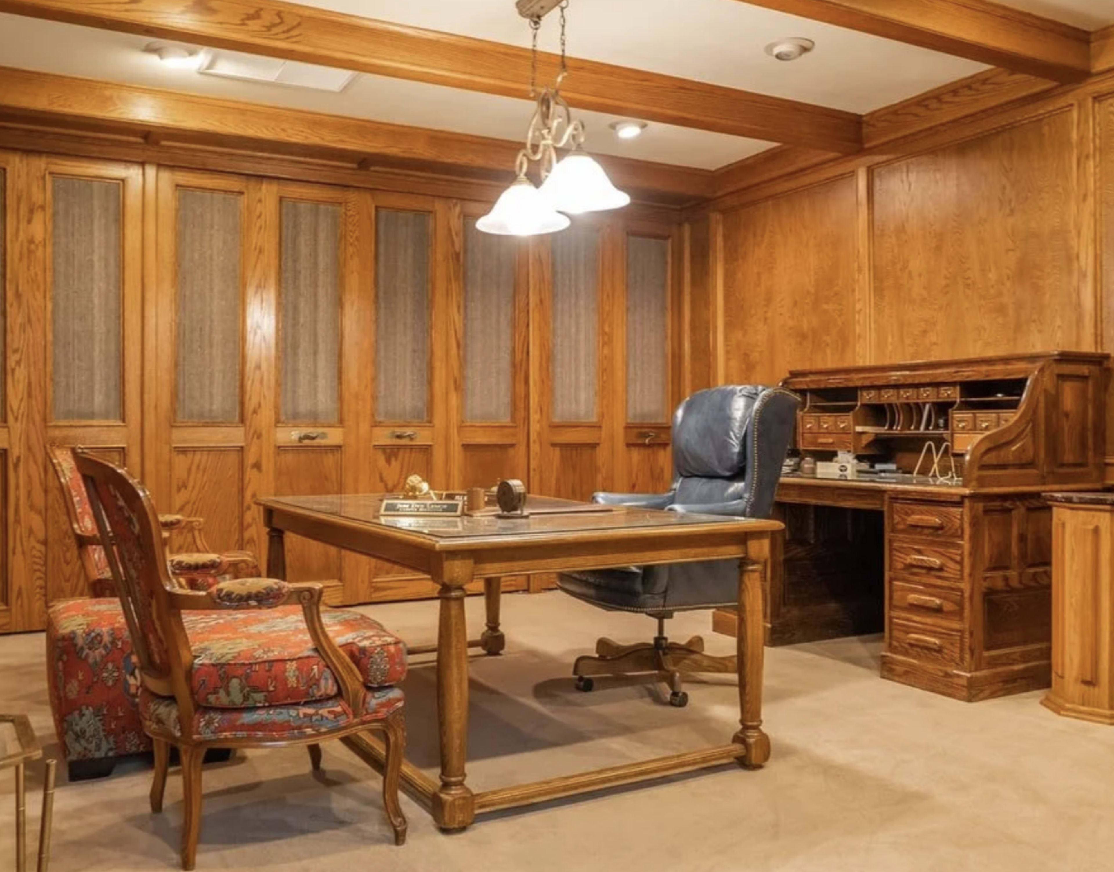 The image shows a wooden office space featuring a large desk, a rolling chair, two additional chairs, and a wooden secretary-style desk against the wall.