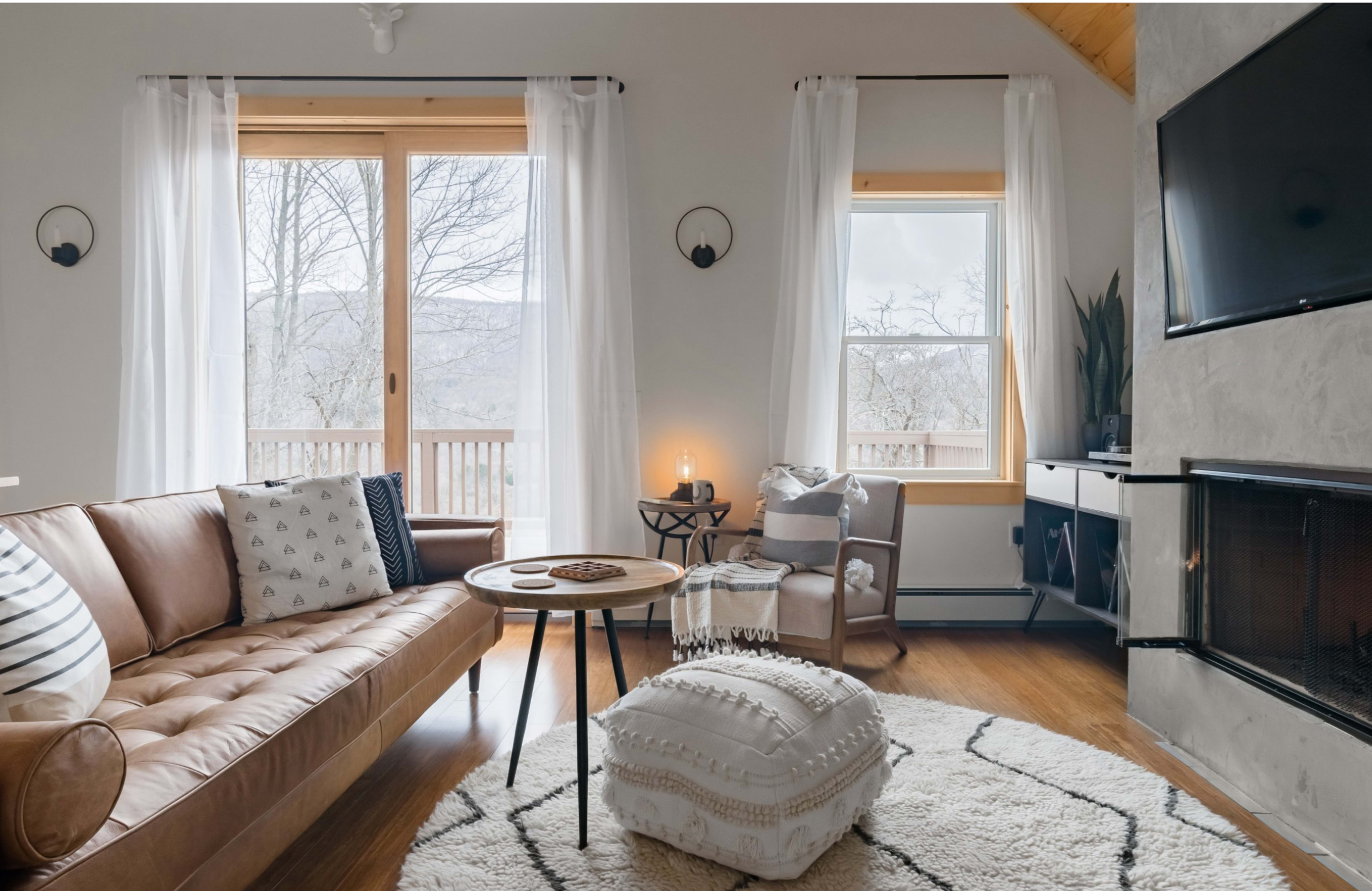 A cozy living room with a brown leather sofa, a round coffee table, and two large windows overlooking a natural landscape.