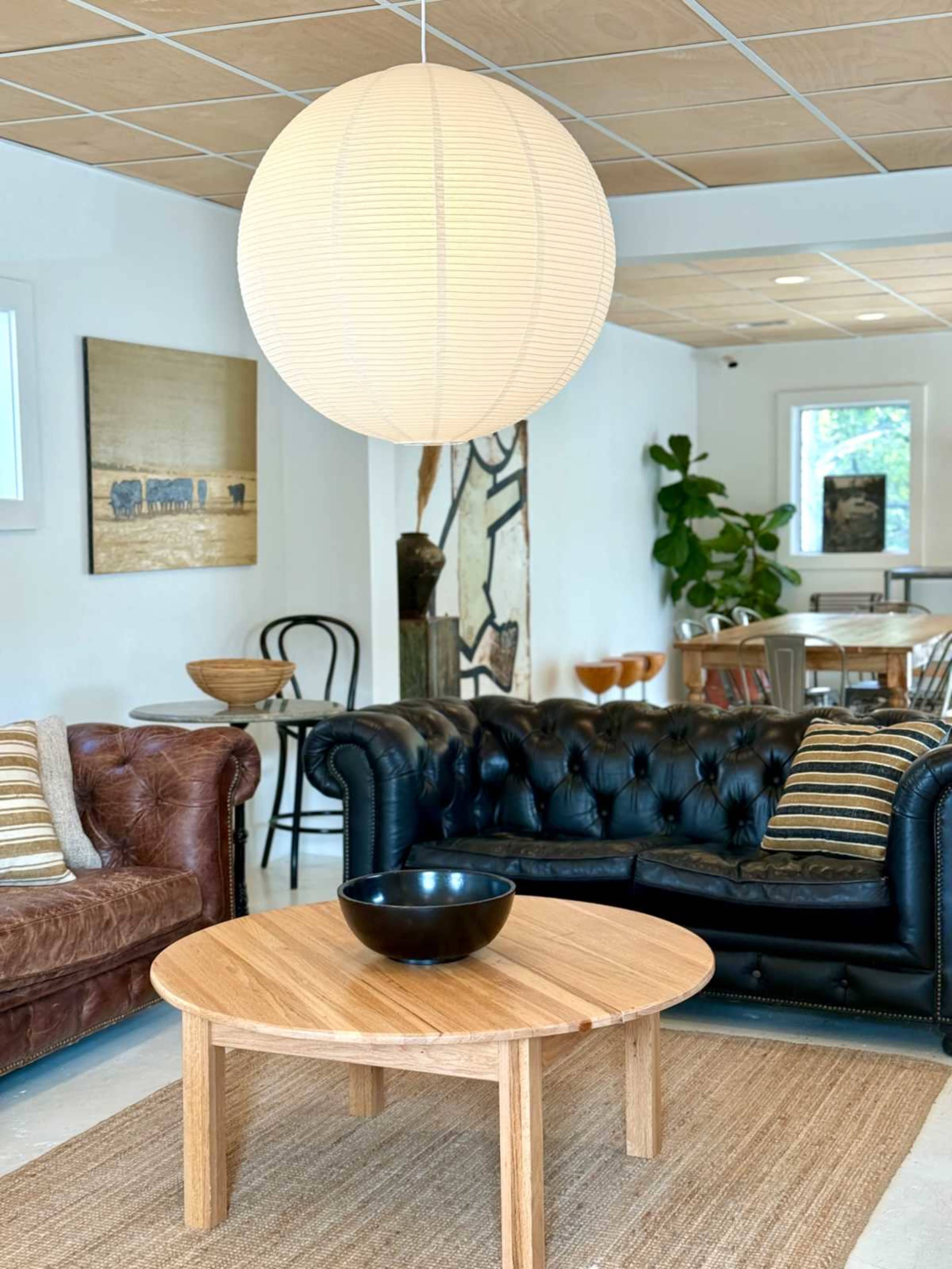 The image shows a living room with a brown leather chesterfield sofa, a black leather chesterfield sofa, a round wooden coffee table, and a large paper lantern hanging overhead, along with various decorative items and plants.
