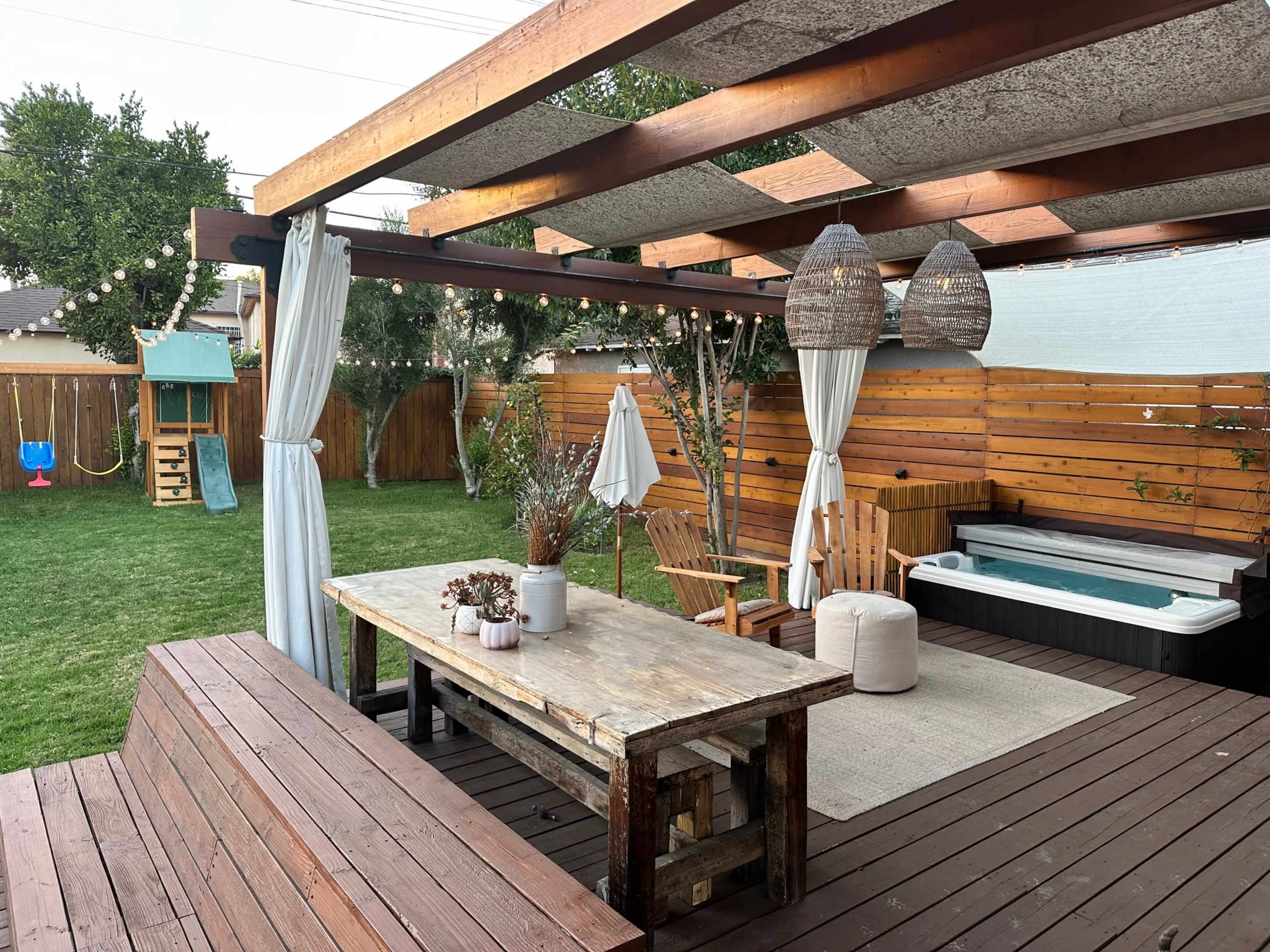A backyard patio with a wooden table, chairs, and a hot tub within a fenced area, surrounded by grass and trees.