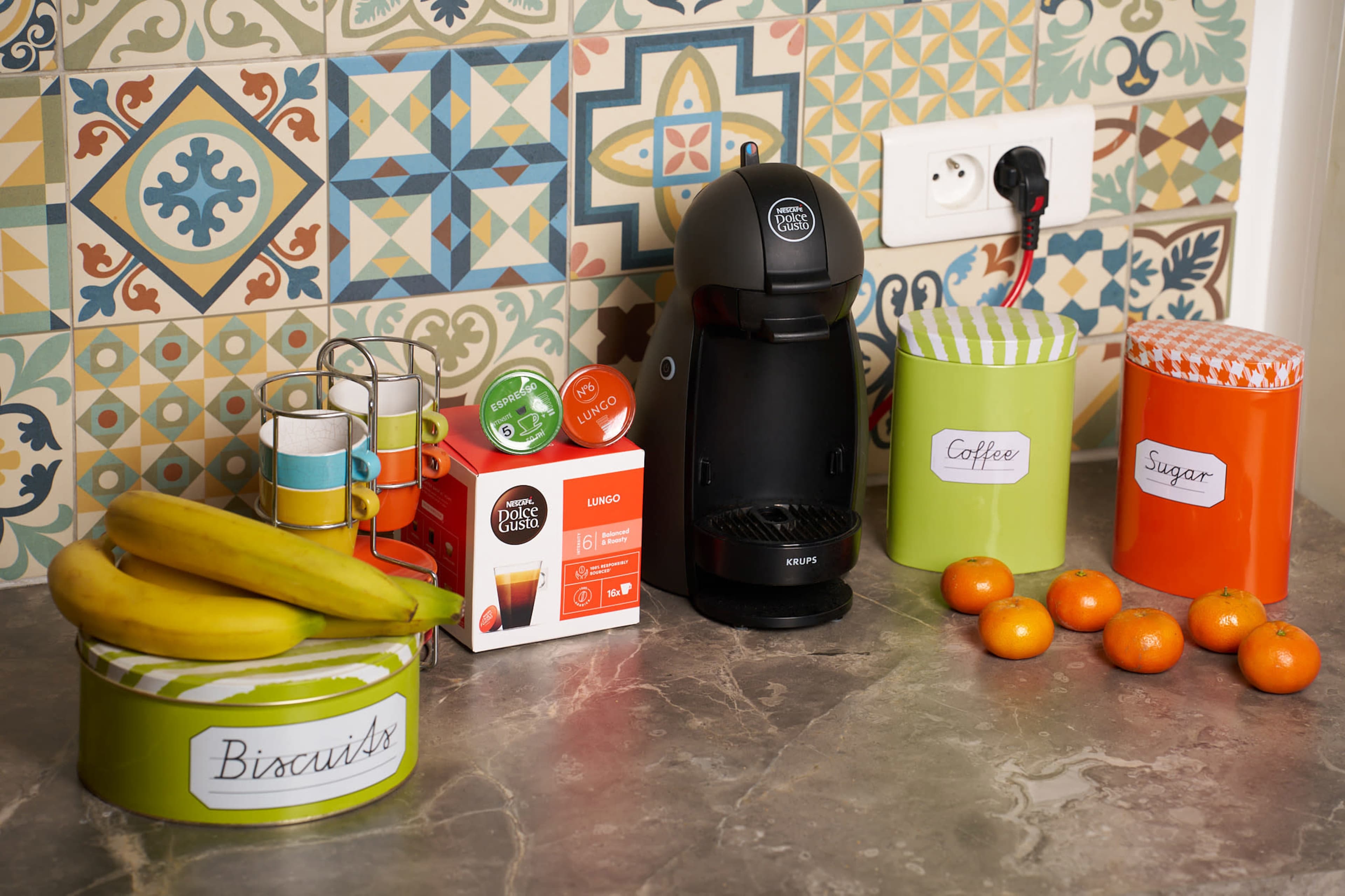 The image shows a kitchen countertop with a coffee machine, a box of coffee pods, a canister labeled "Coffee," another labeled "Sugar," a green canister, bananas, and a bowl of oranges, all set against a colorful tiled backsplash.