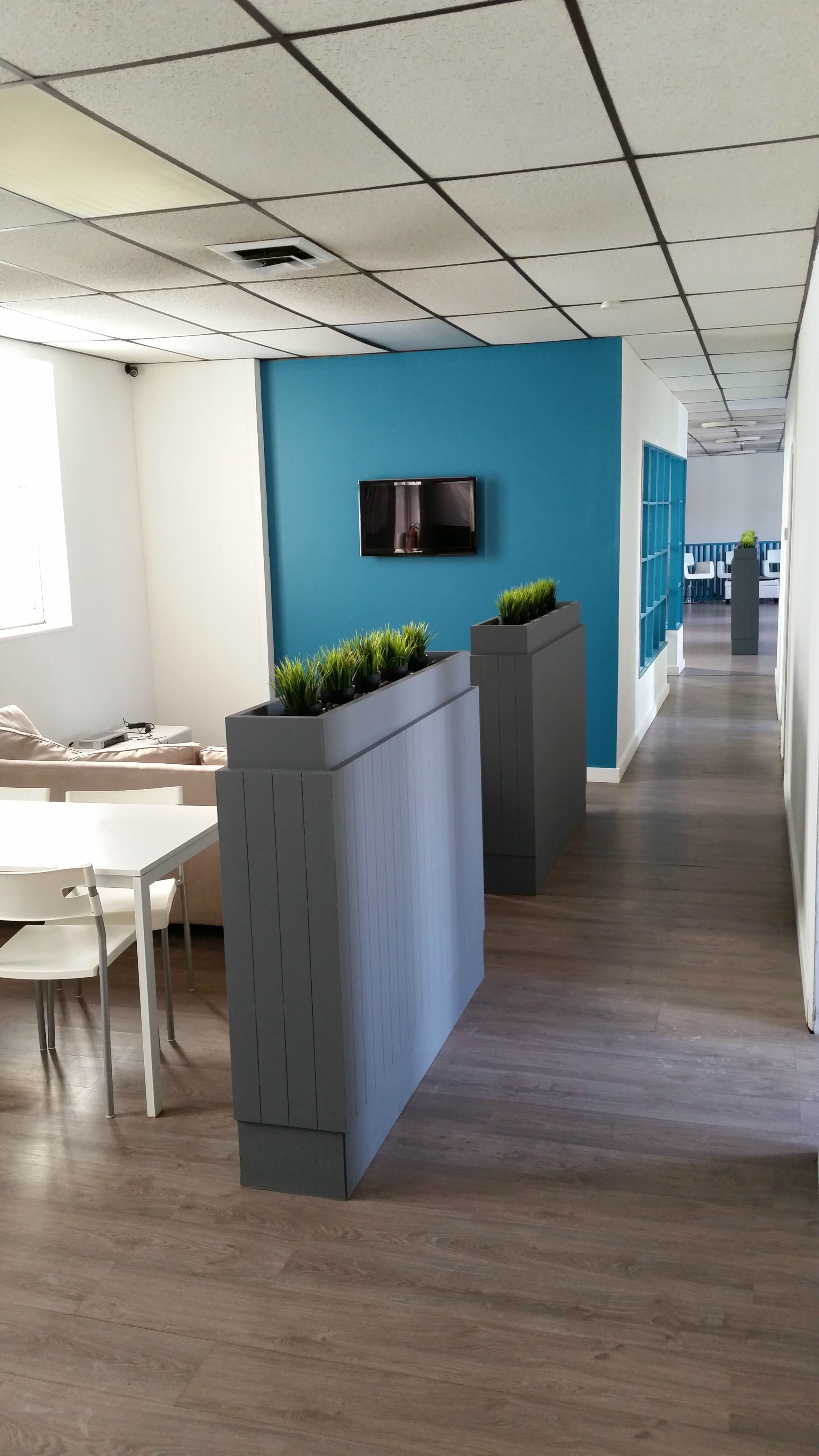 A modern office interior with a long hallway featuring potted plants, a wall-mounted TV, and a seating area at one end.