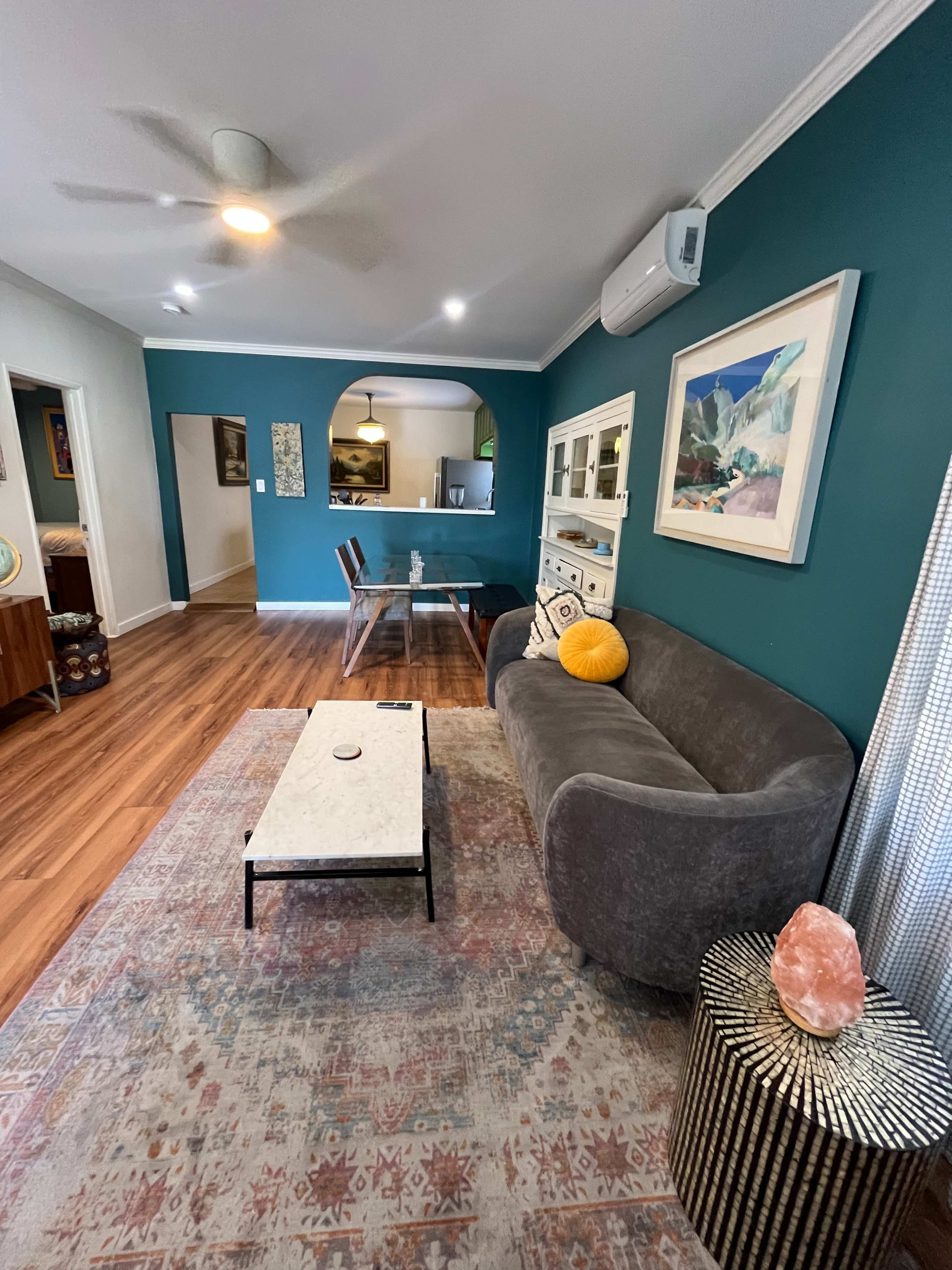 The image shows a well-decorated living room featuring a gray sofa, a coffee table, and a colorful rug, with teal walls and various decorative elements.