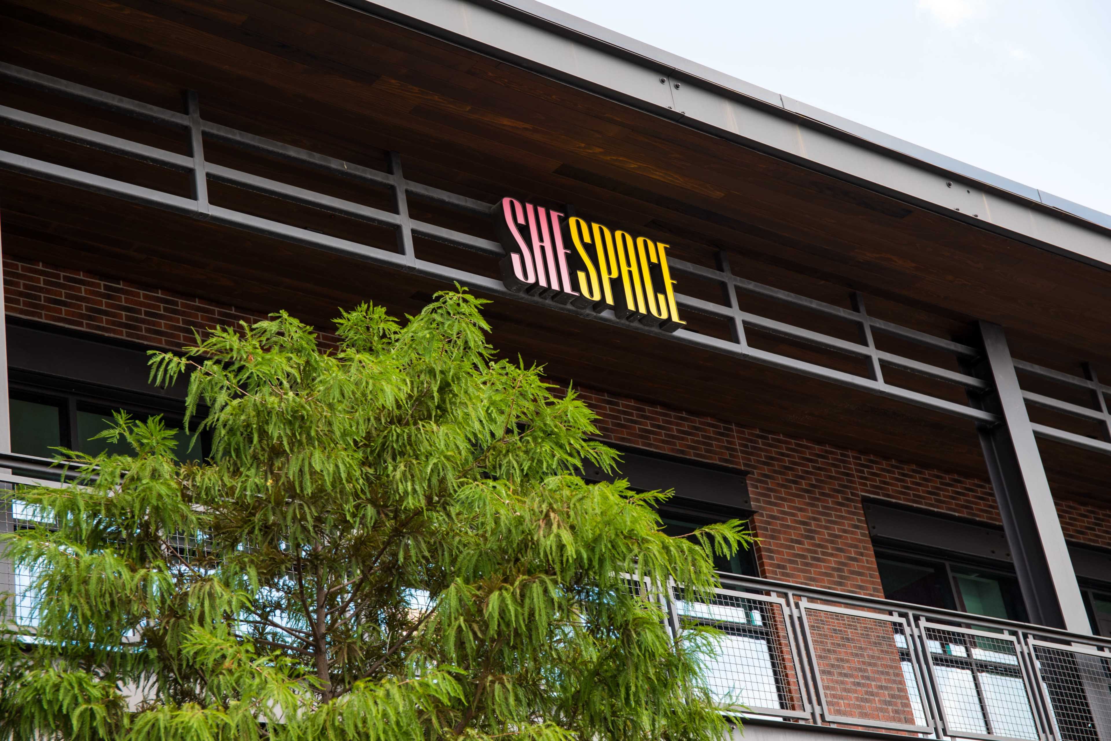 A building features a sign that reads "SHE SPACE" above a green tree.