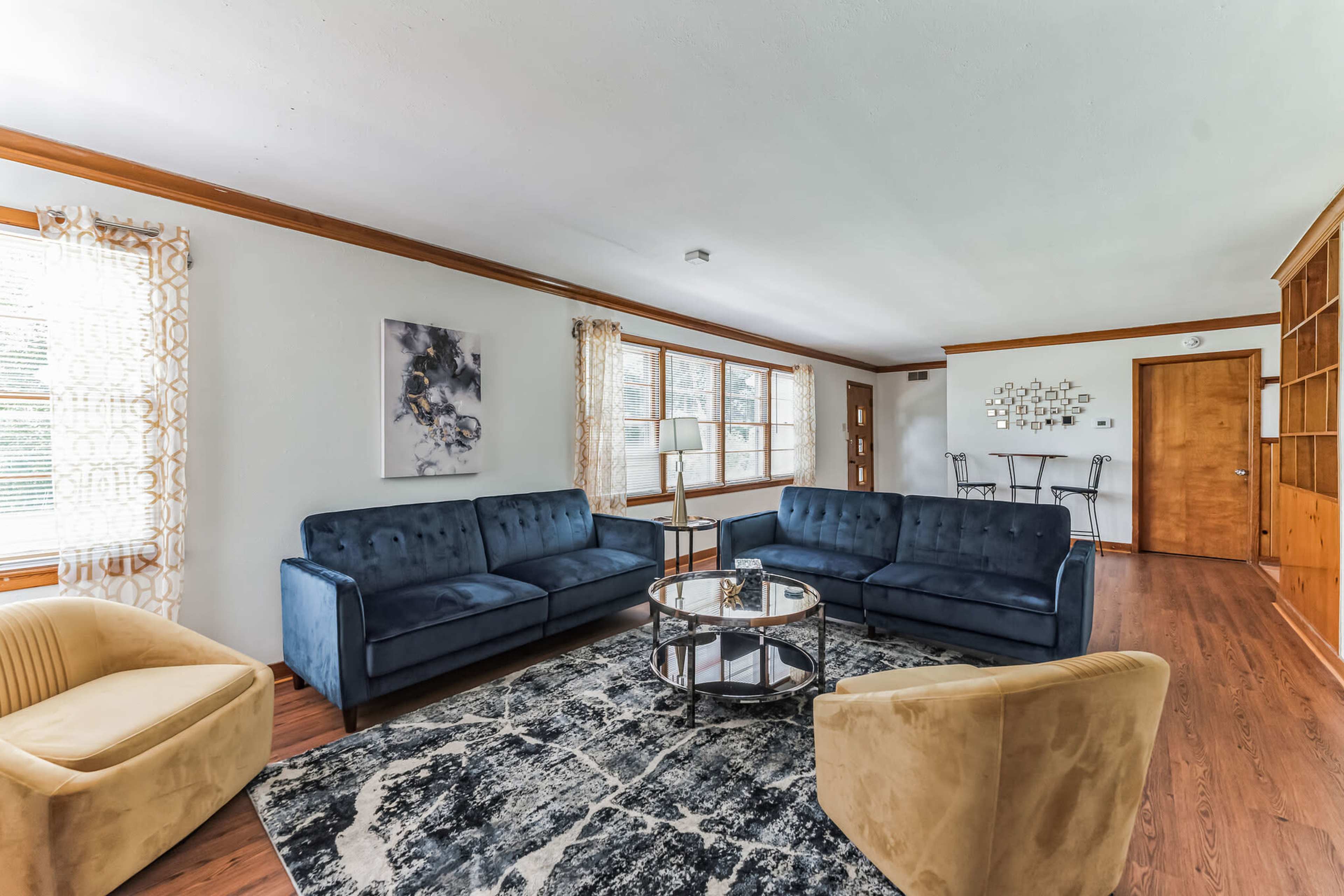 The living room features two navy blue sofas, one beige armchair, a round glass coffee table, and large windows with patterned curtains.