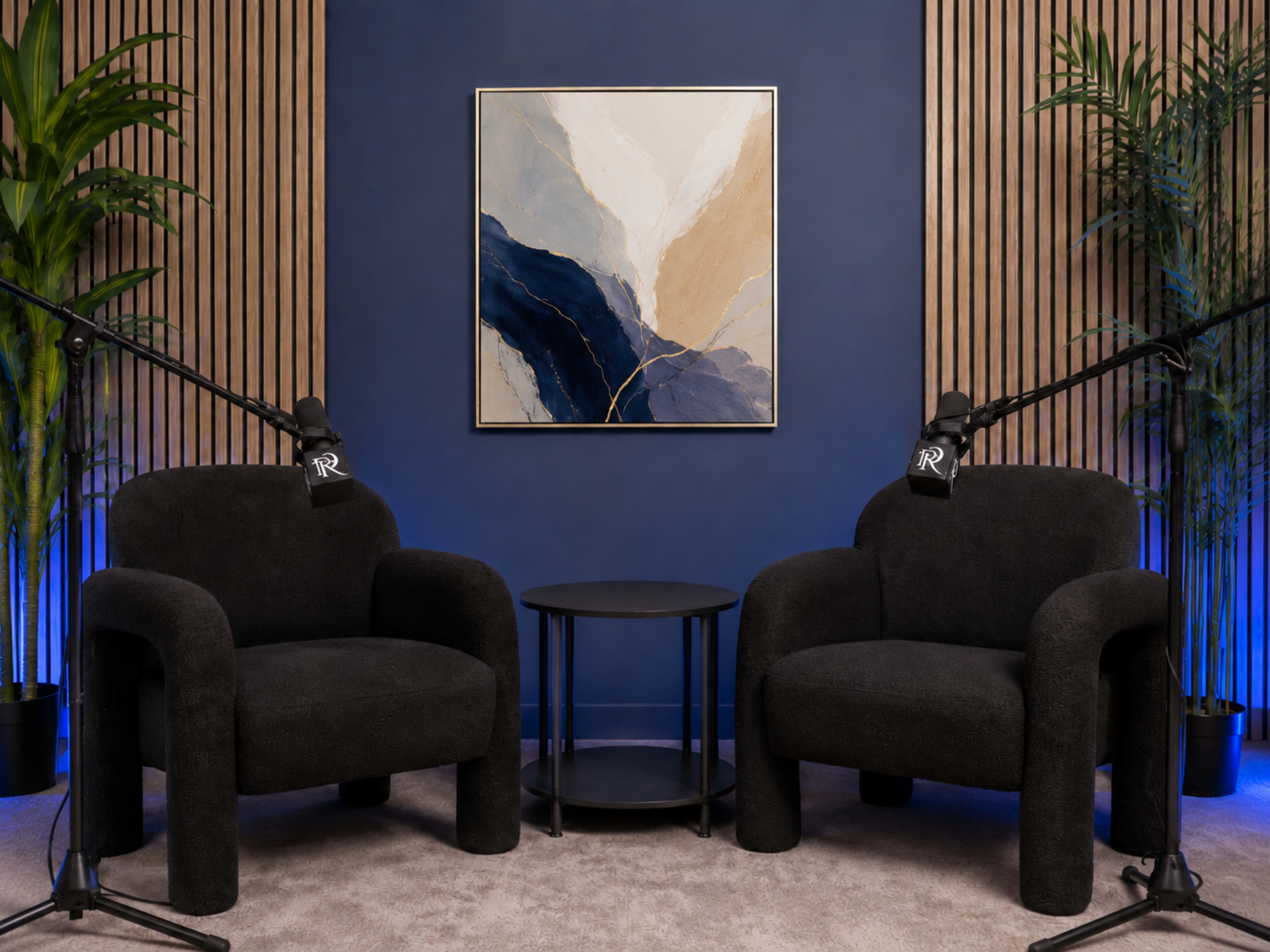 The image shows a modern seating area with two black, rounded armchairs flanking a small black table, set against a blue wall and adorned with a large abstract painting above.