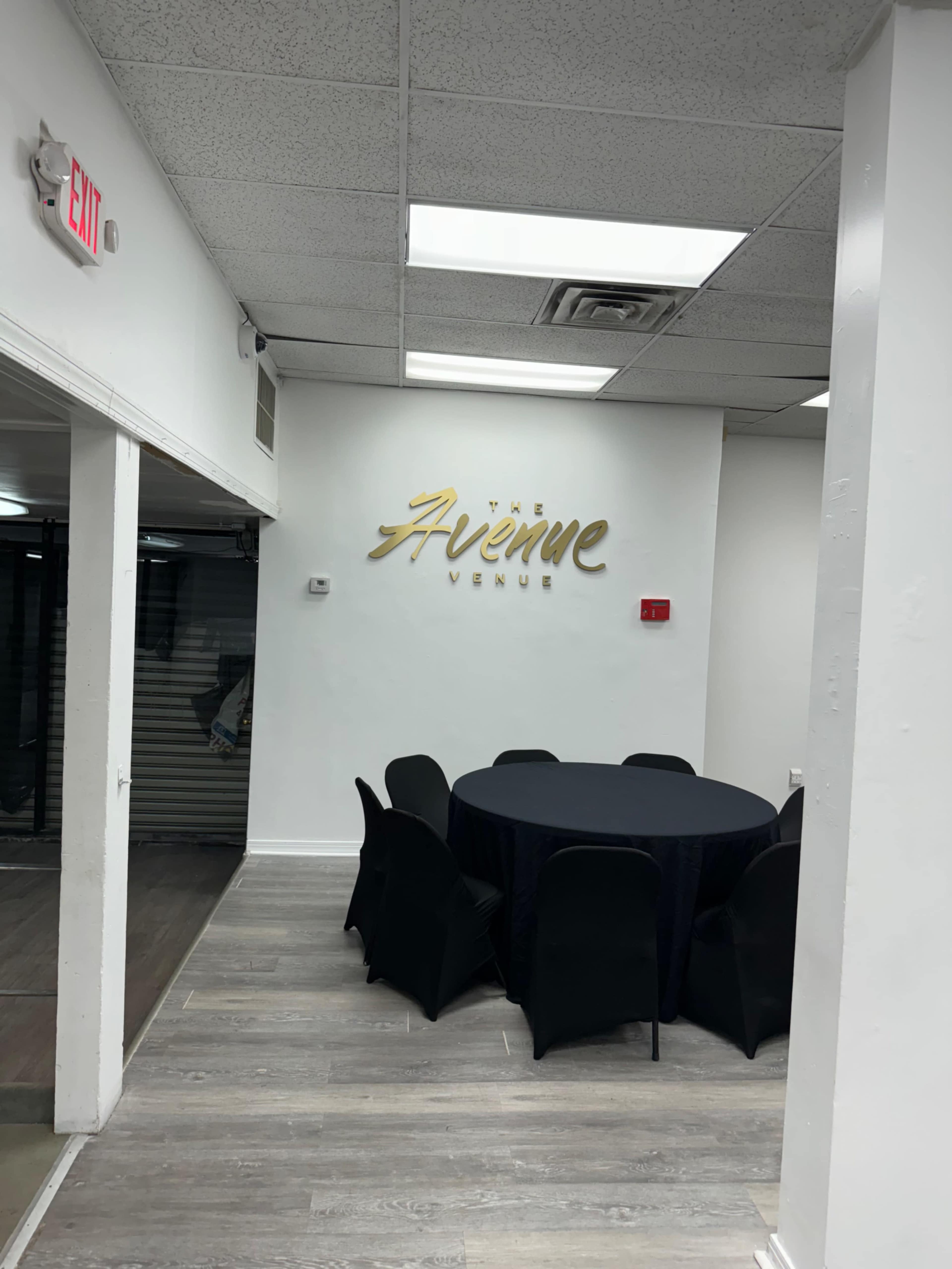 A spacious indoor venue with a circular black table surrounded by black chairs, and a gold sign on the wall reading "The Avenue Venue."