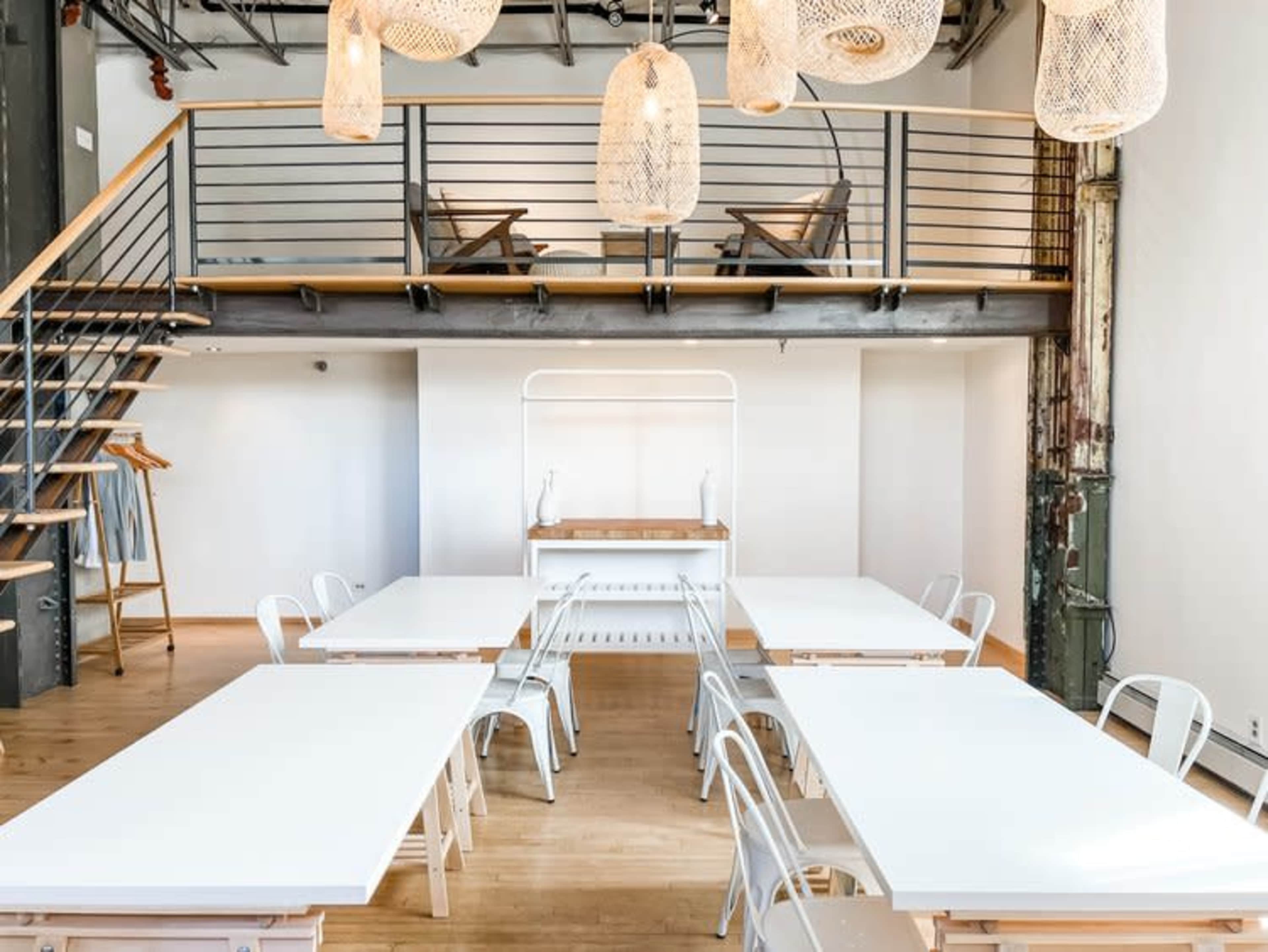 A modern interior space with several white tables arranged in a large area under hanging pendant lights, featuring a staircase leading to a balcony.