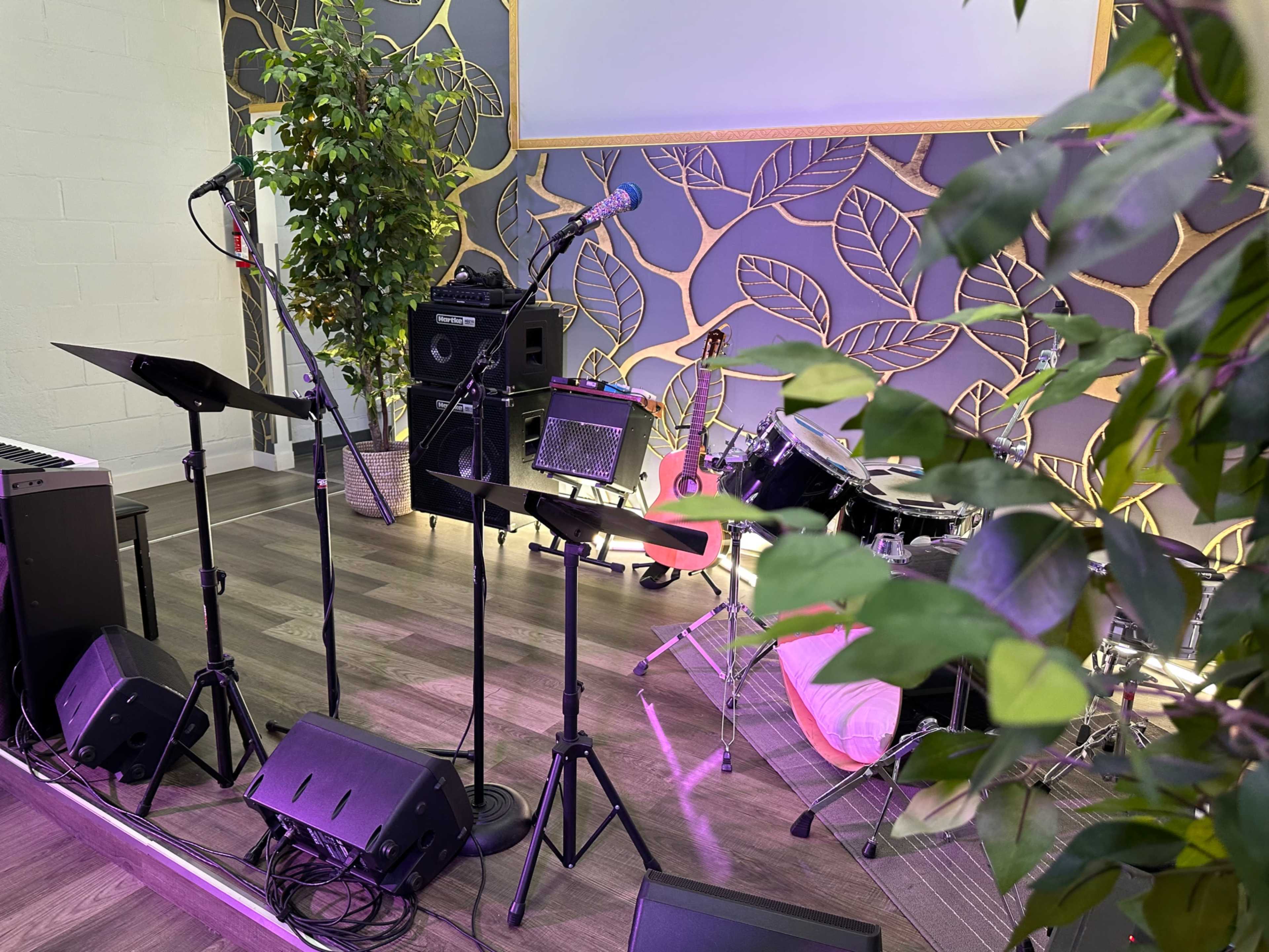 The image shows a stage setup with musical instruments, including a keyboard, microphones, and a drum kit, surrounded by decorative plants.
