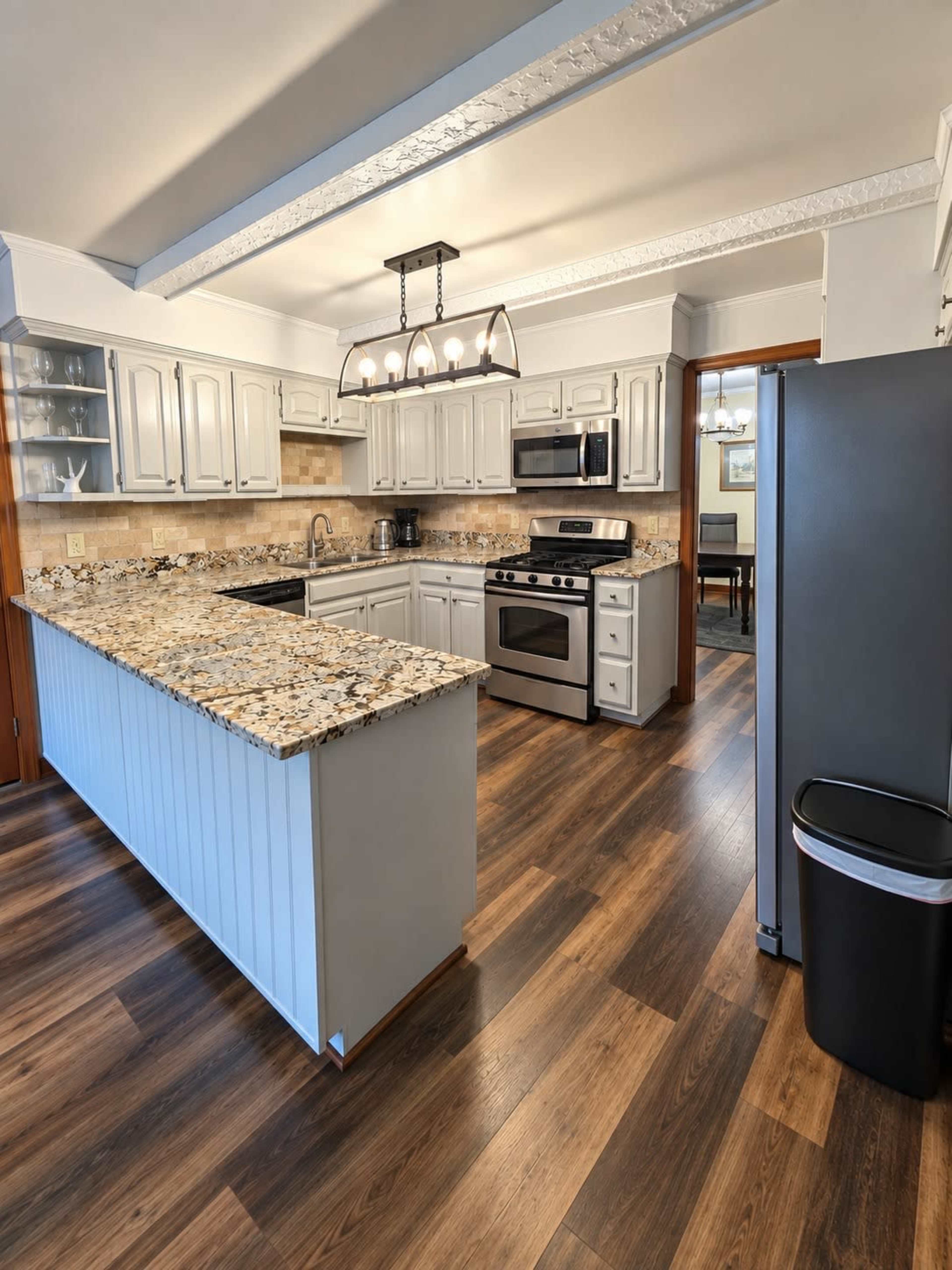 A modern kitchen features gray cabinetry, stainless steel appliances, a large granite countertop island, and wooden flooring.