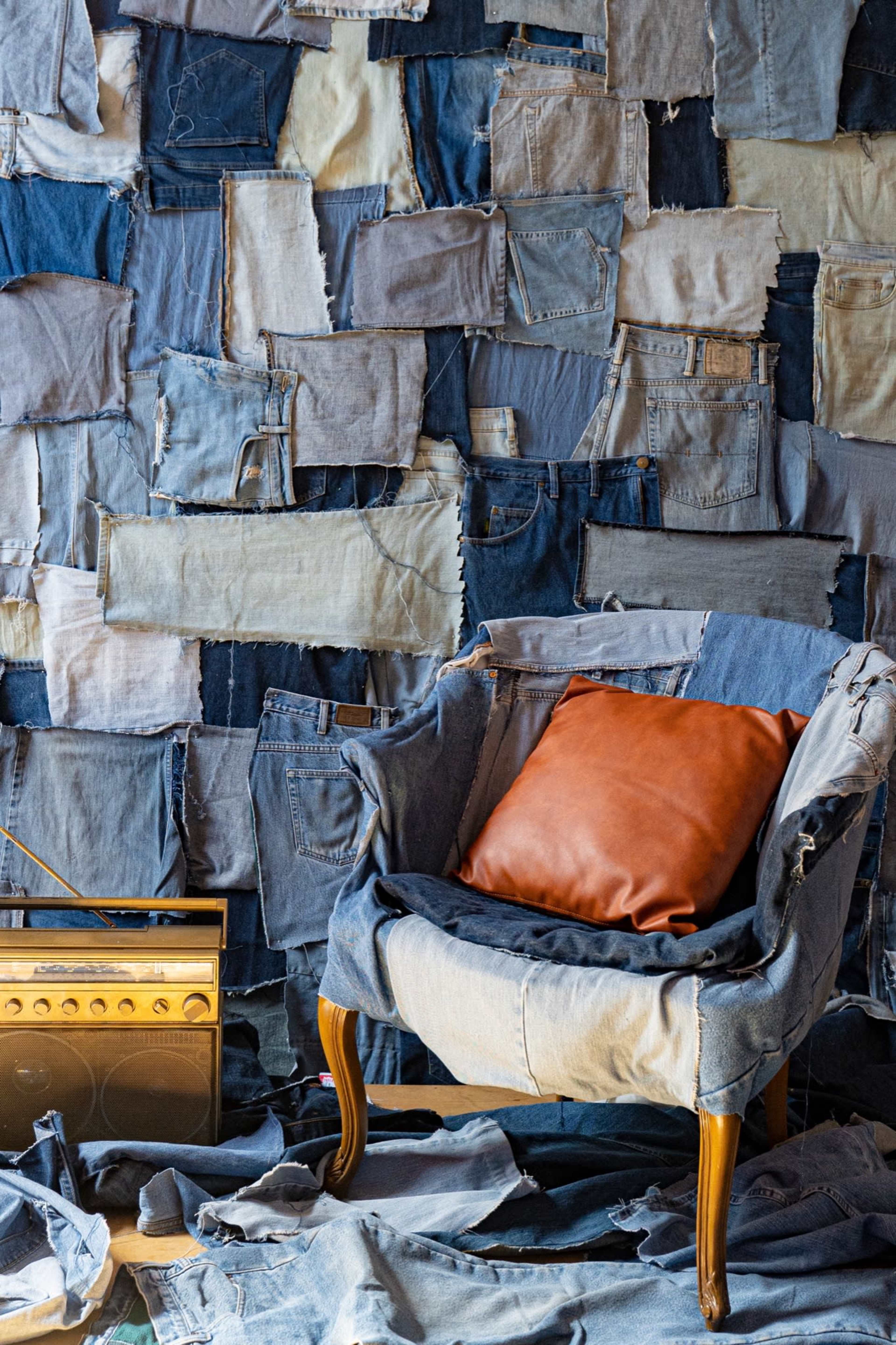 A chair with a brown pillow is positioned in front of a wall covered in various pieces of denim fabric, alongside a vintage radio on the floor.