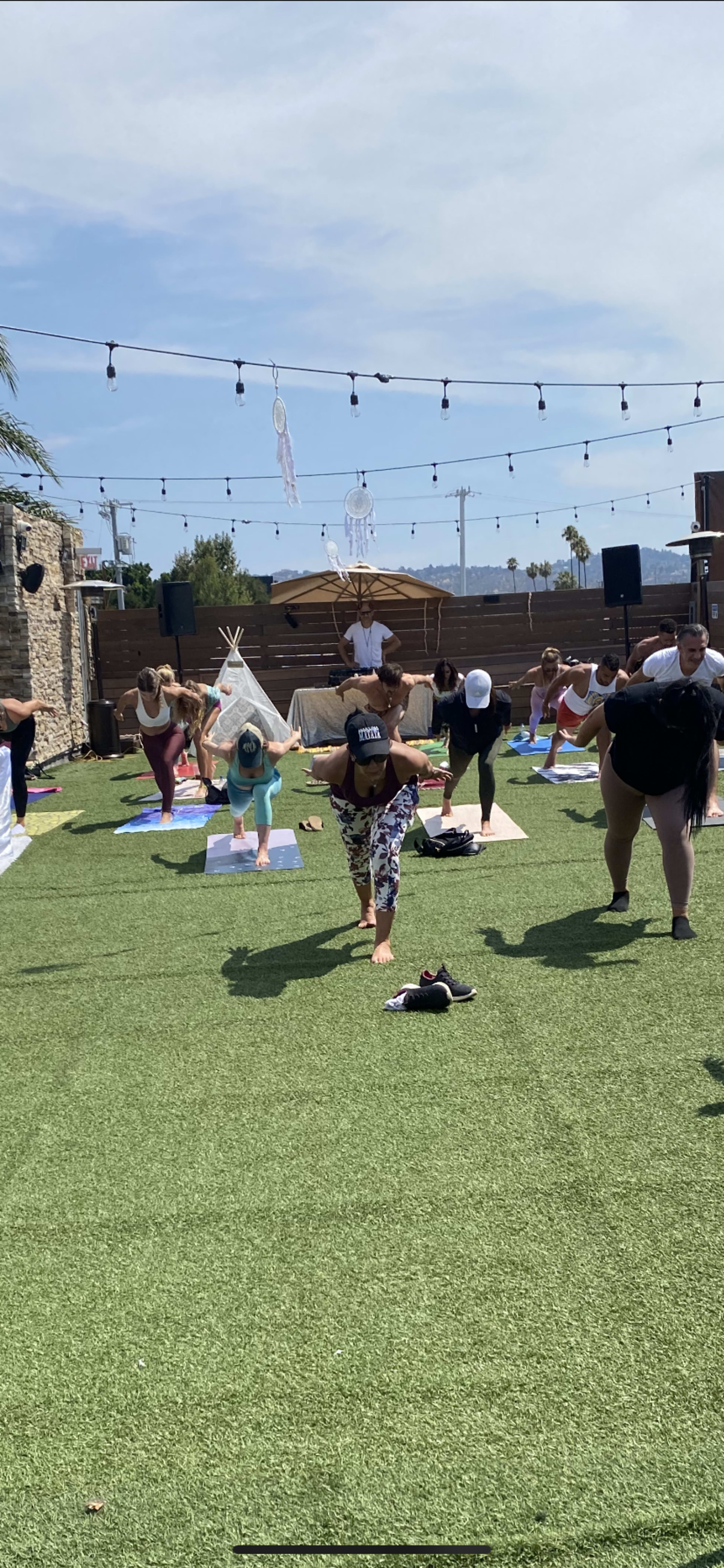A group of people is practicing yoga on mats on a green lawn under a clear sky, with decorative lights hanging overhead.
