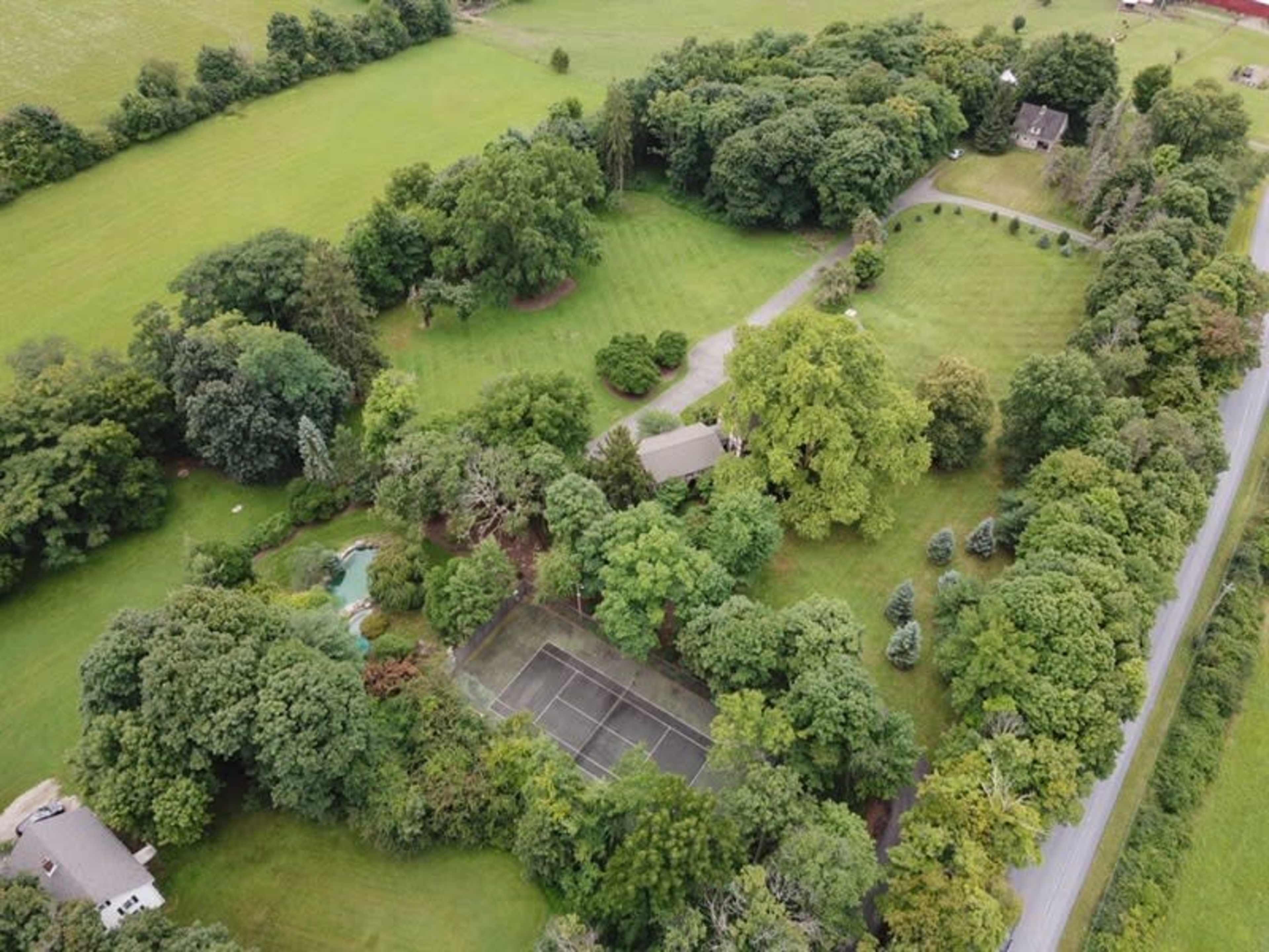 An aerial view of a large, green area featuring several trees, a house, and a tennis court surrounded by dense foliage.