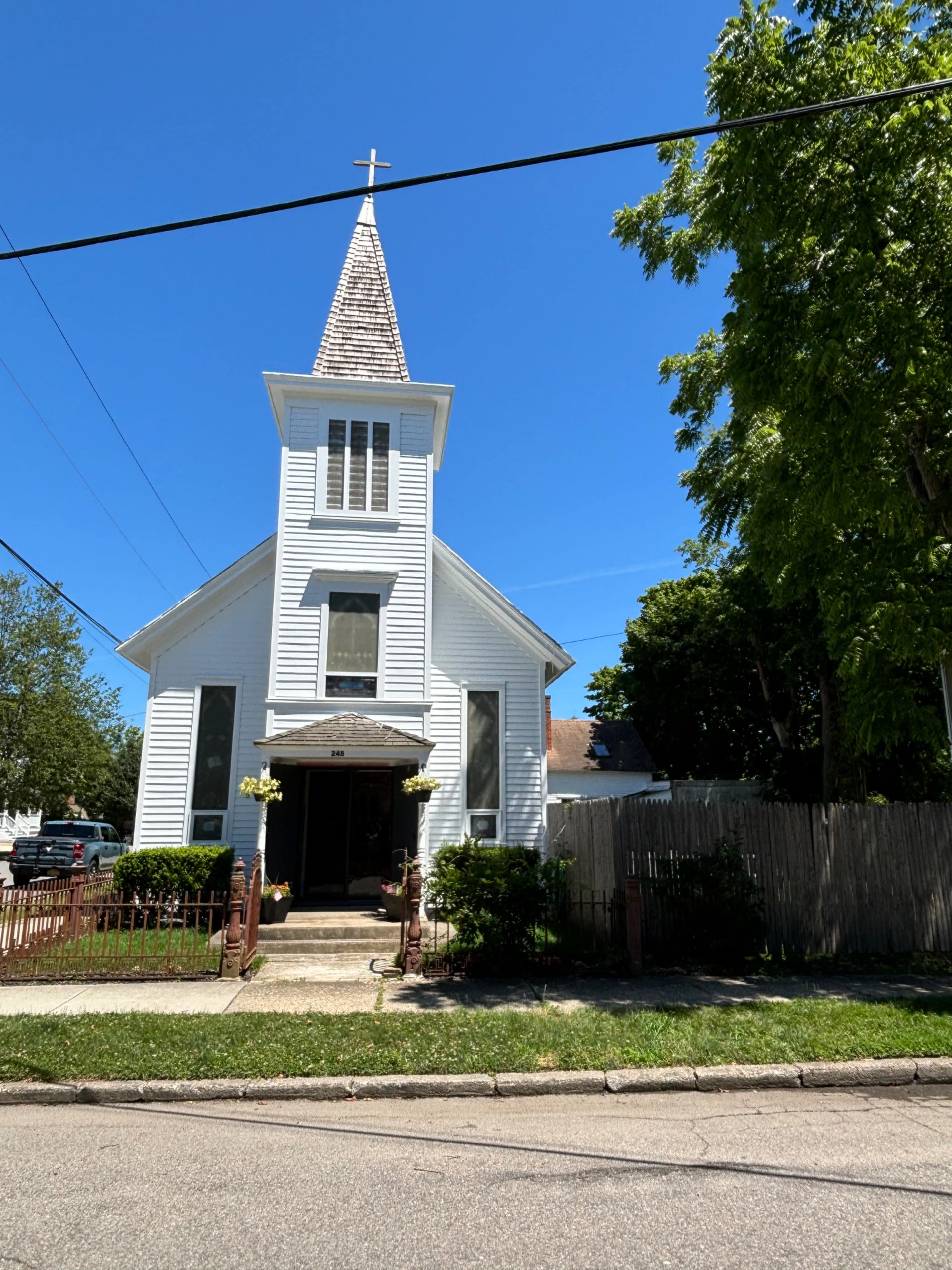 A white, wooden church with a tall steeple and cross sits at the corner of a street, surrounded by greenery.
