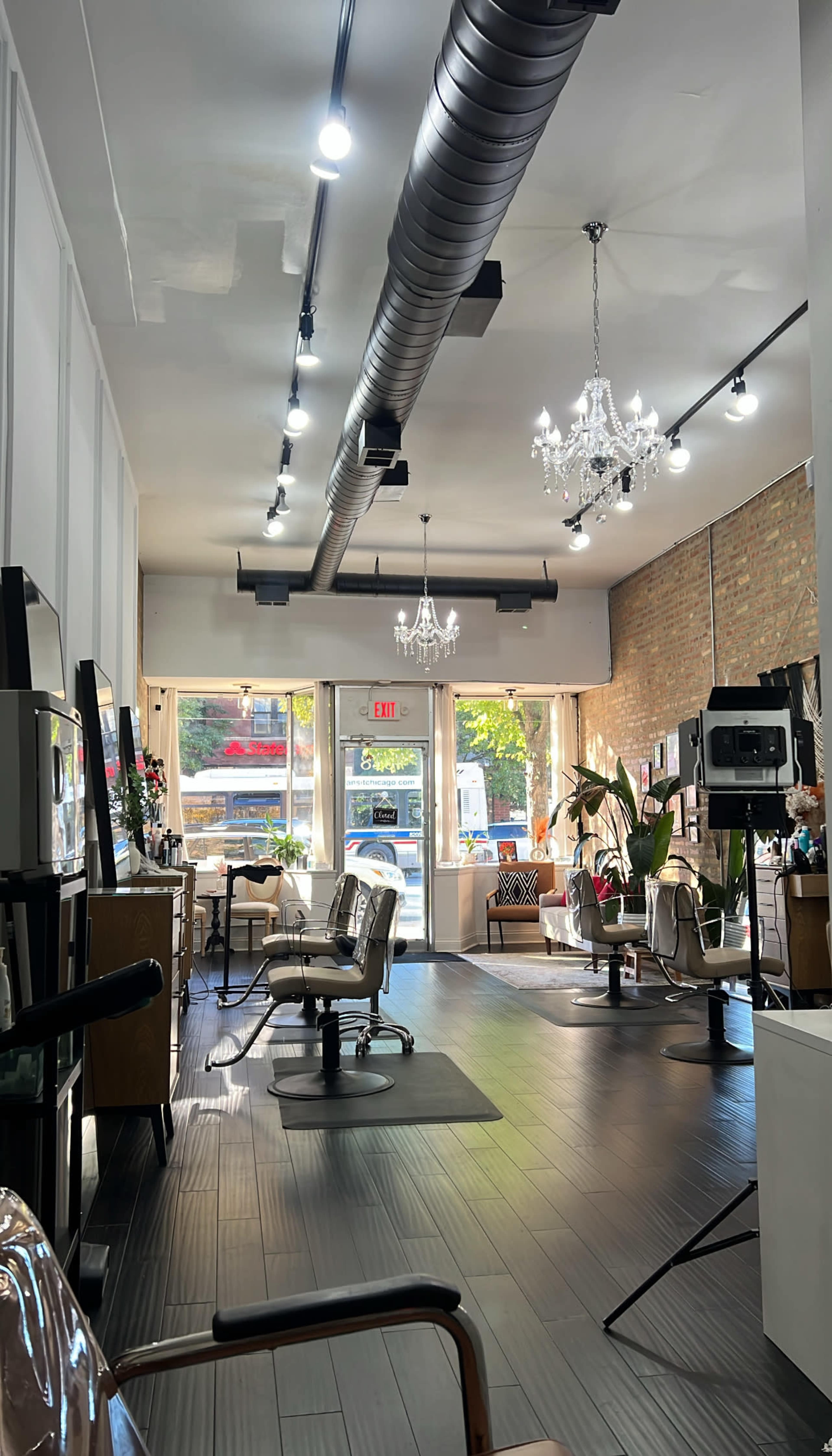 The image shows a bright hair salon with several styling stations, large windows letting in natural light, and decorative chandeliers hanging from the ceiling.