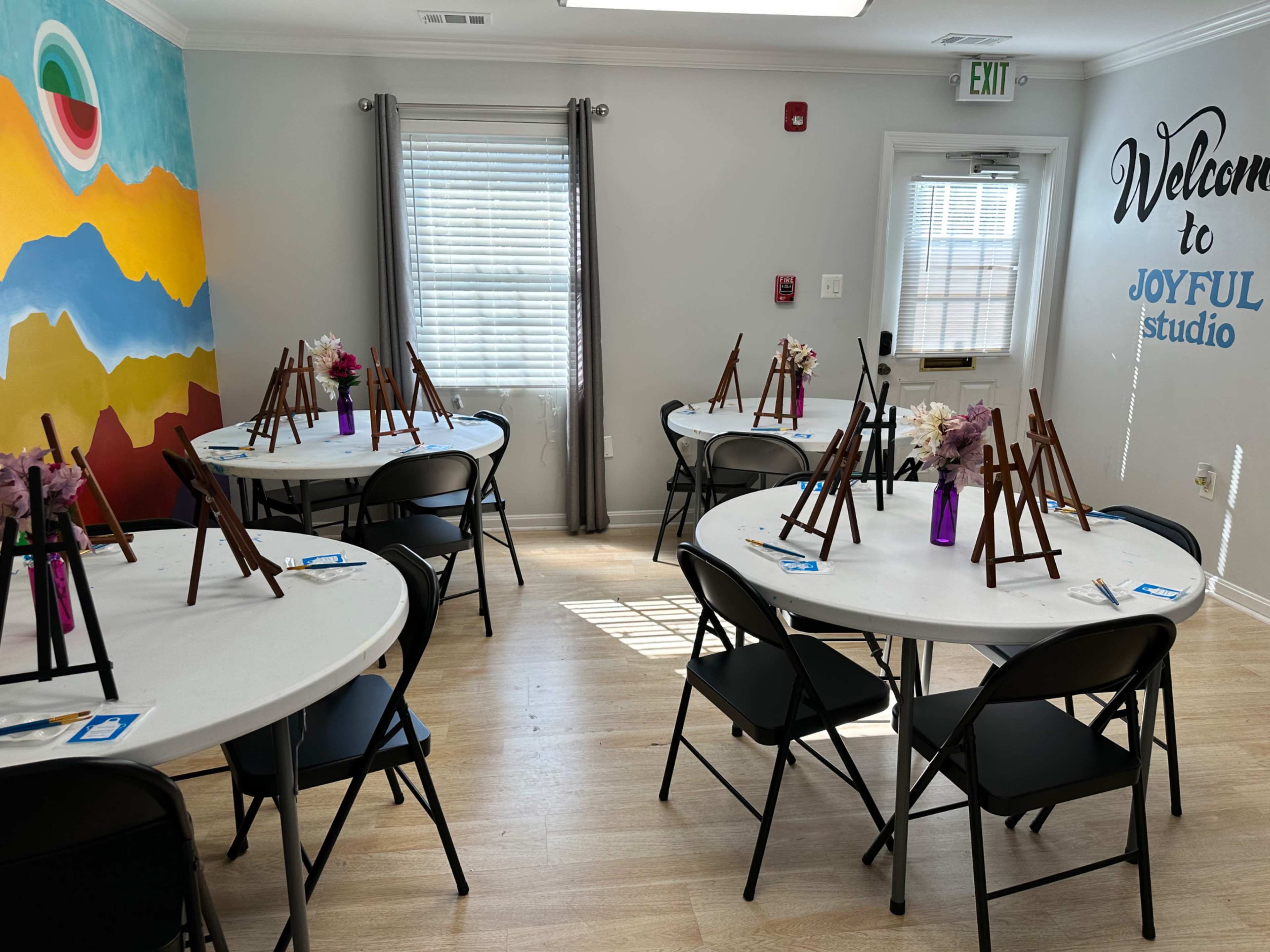 A brightly lit classroom with several round tables set up, each equipped with an easel and a small vase of flowers, creating a workshop layout.