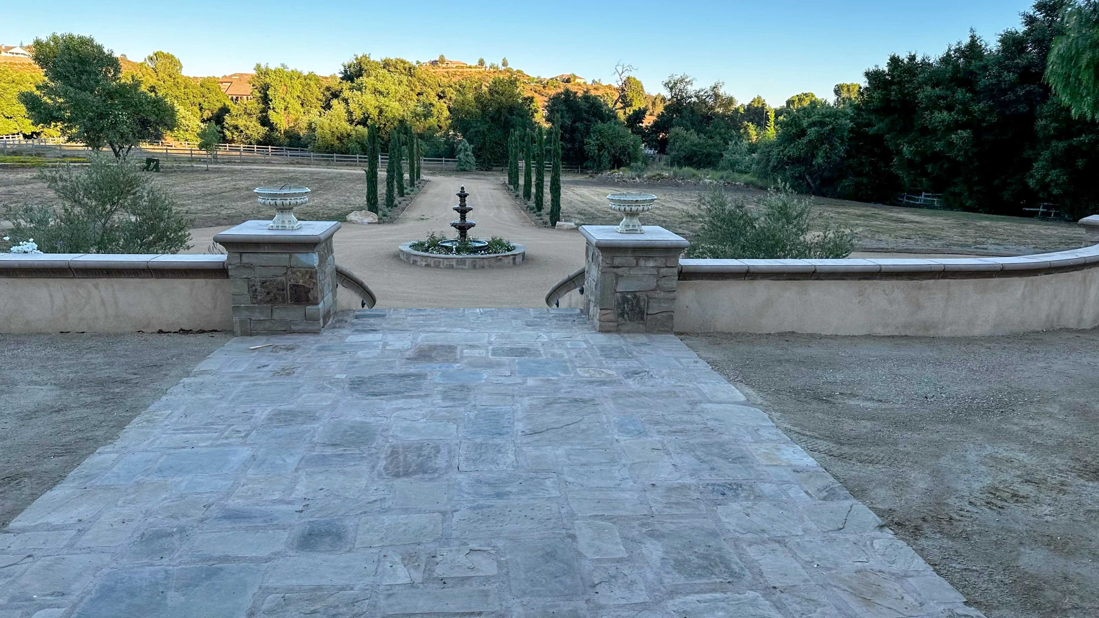 A stone pathway leads to a circular garden with a fountain, surrounded by greenery and lined with tall cypress trees.