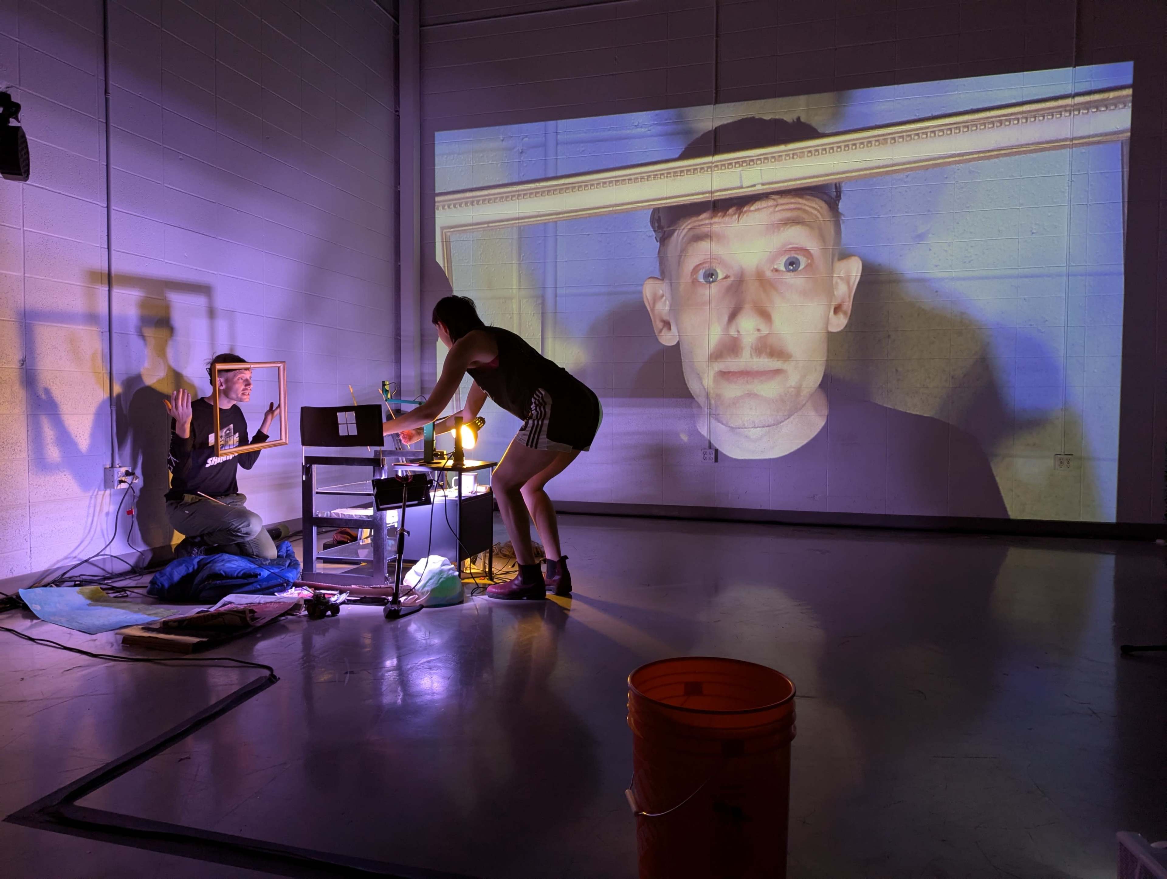 A woman adjusts equipment while a man appears projected inside a picture frame on a wall in a dimly lit studio.