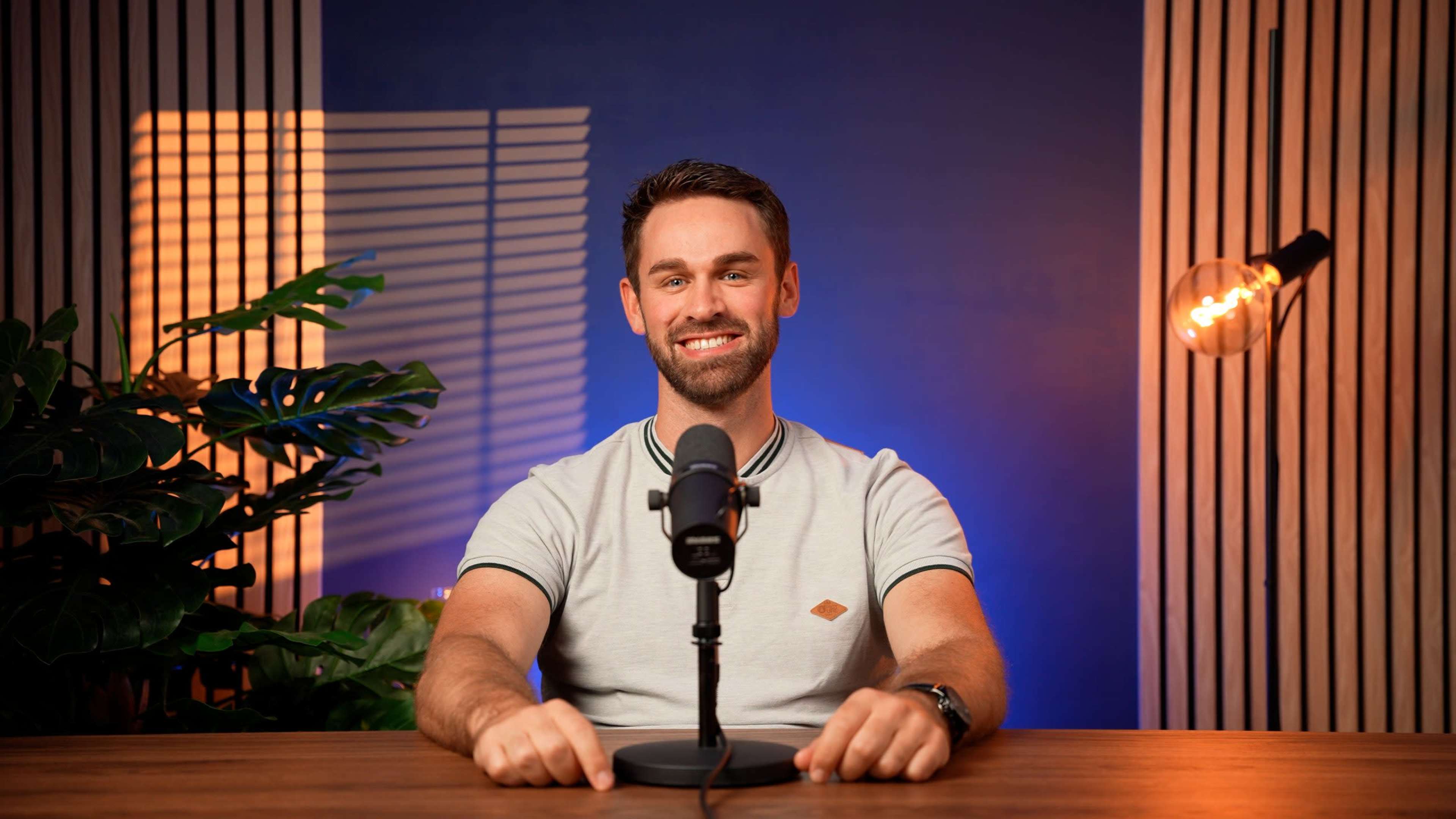 A man sits at a wooden table in front of a microphone, with plants and decorative lighting in the background.