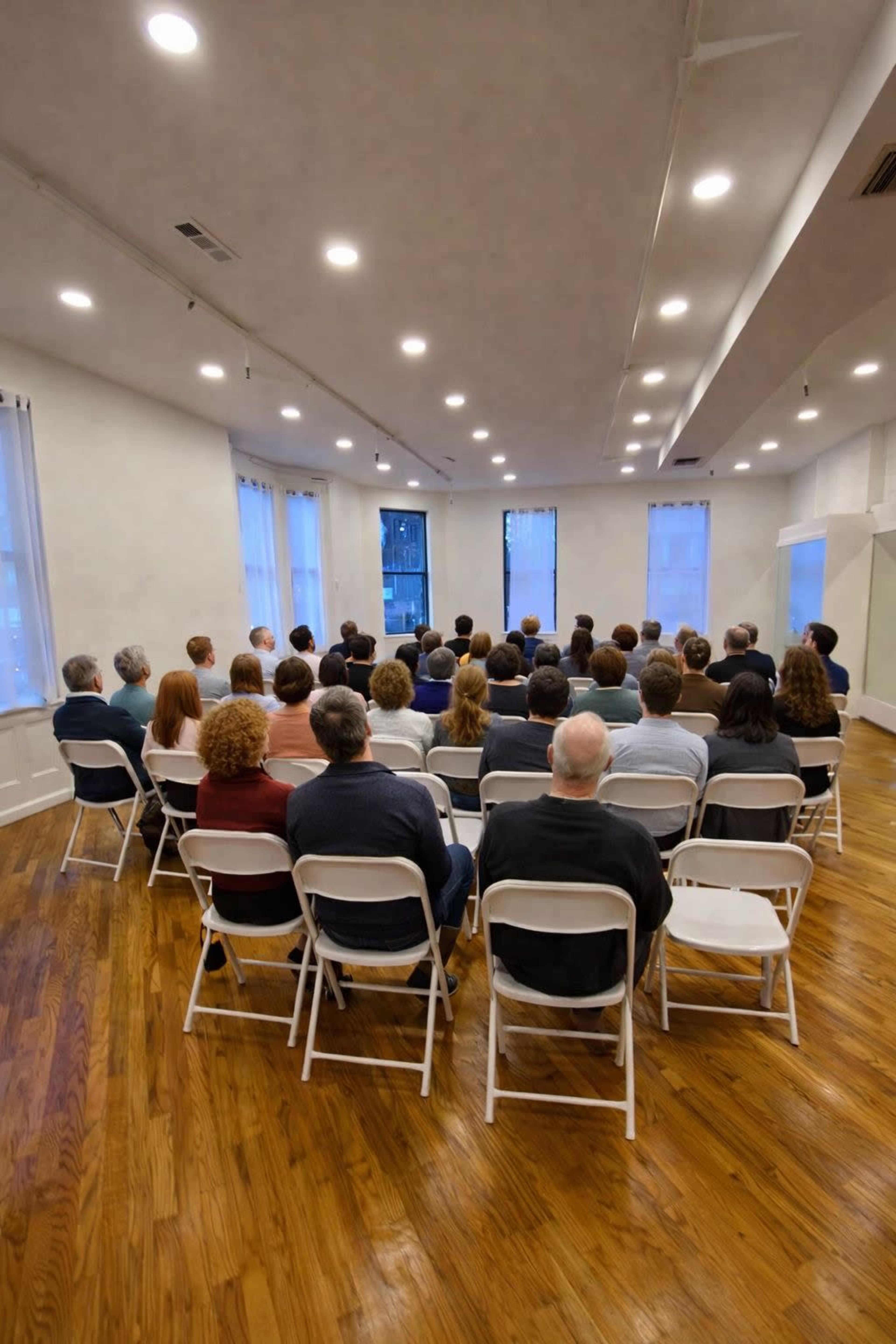 A group of people sits in rows facing a speaker at the front of a well-lit room.