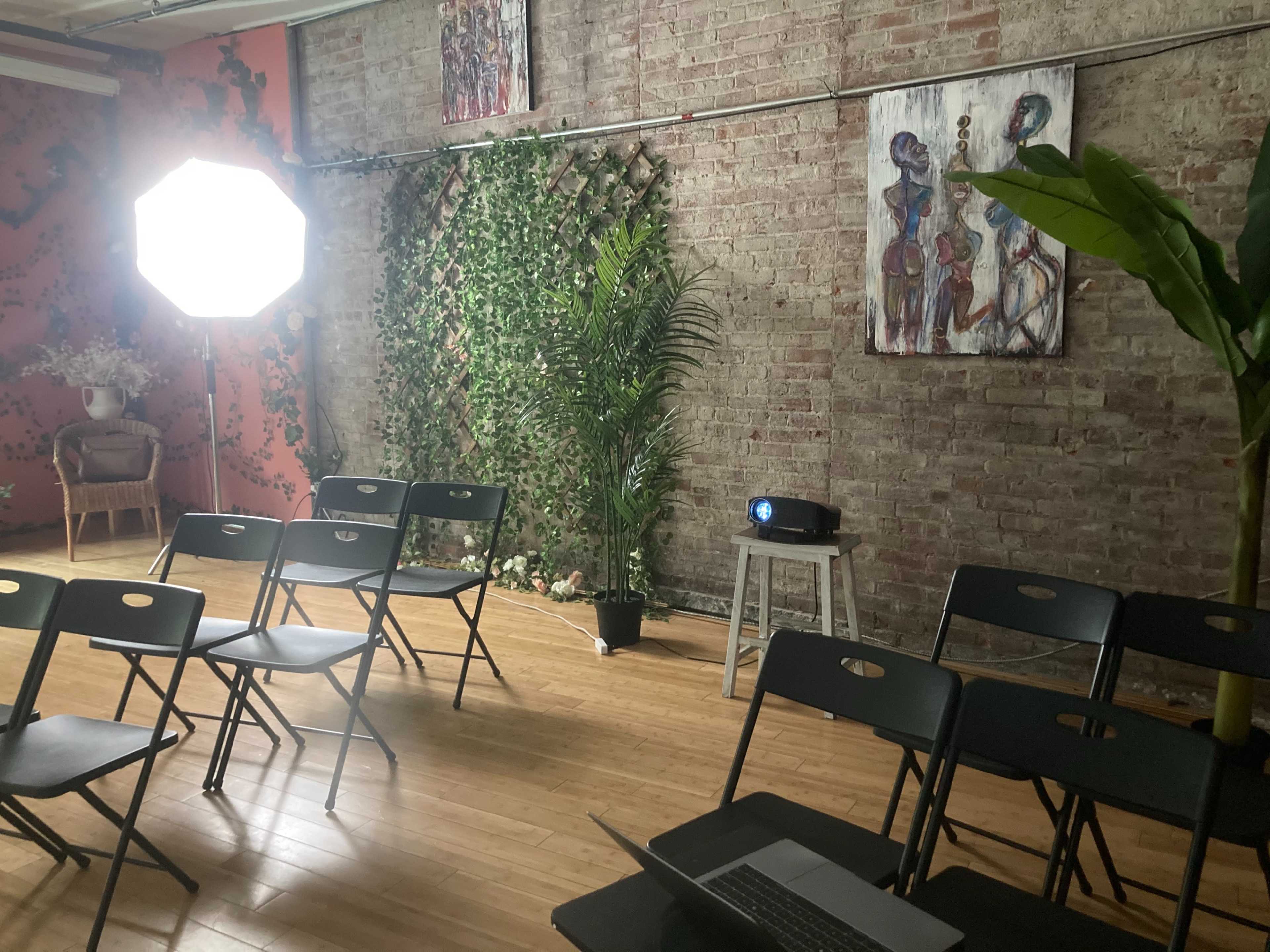 A well-lit room set up for a presentation, featuring black folding chairs arranged in rows, a projector on the floor, and a large light source next to a green plant and artwork on the brick walls.