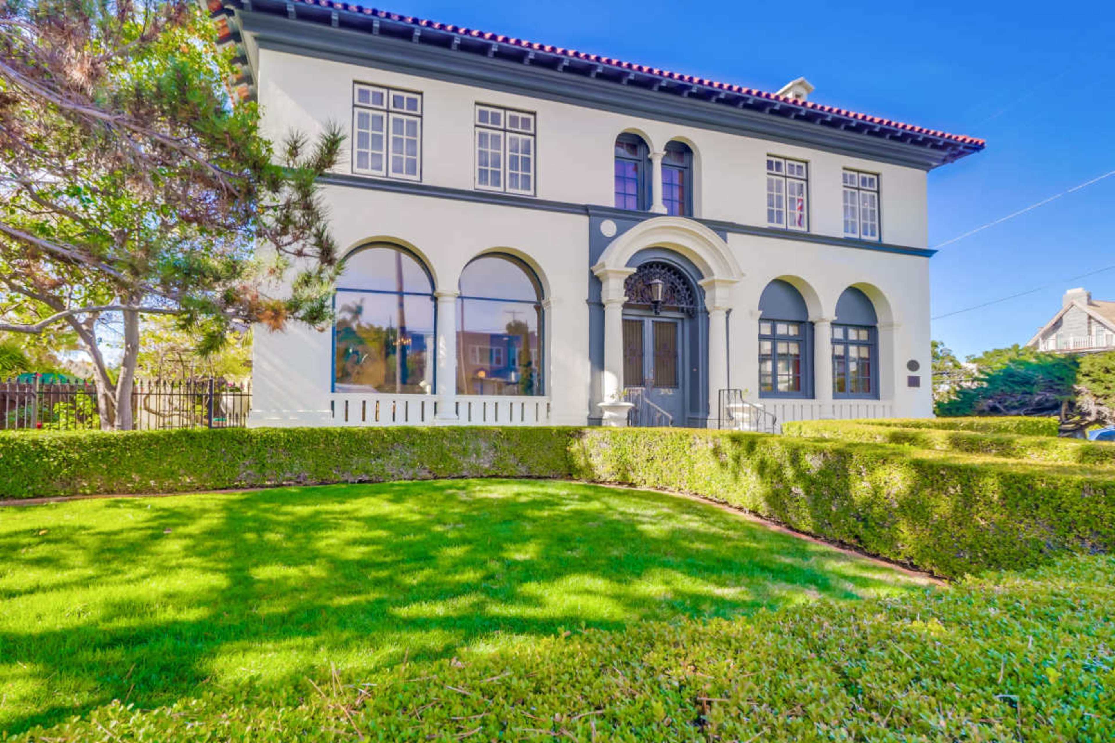The image depicts a large, two-story white building with arched windows and a manicured lawn surrounded by a neatly trimmed hedge.