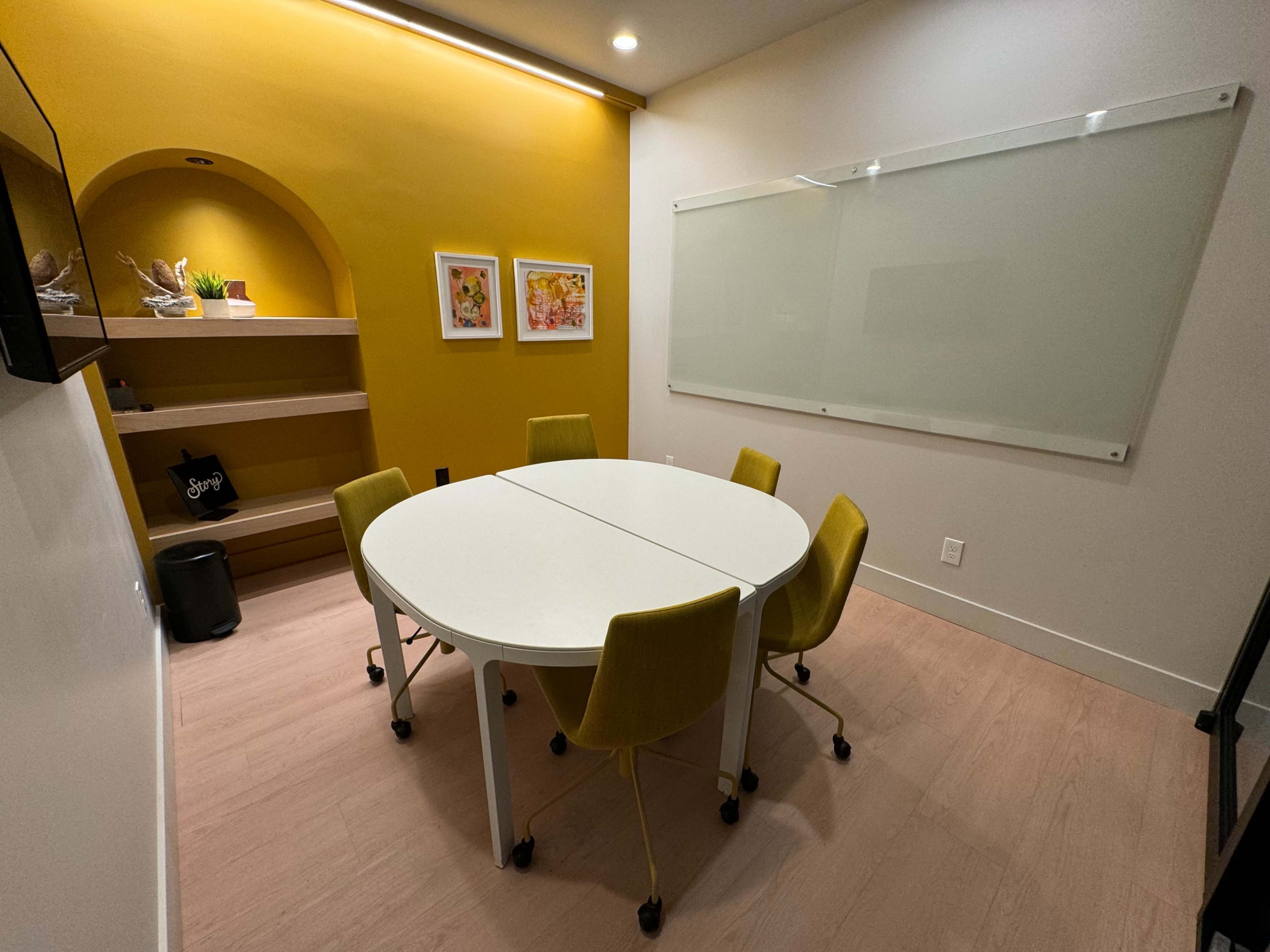 A small meeting room features a round white table surrounded by four yellow chairs, with a light yellow wall and a glass whiteboard.