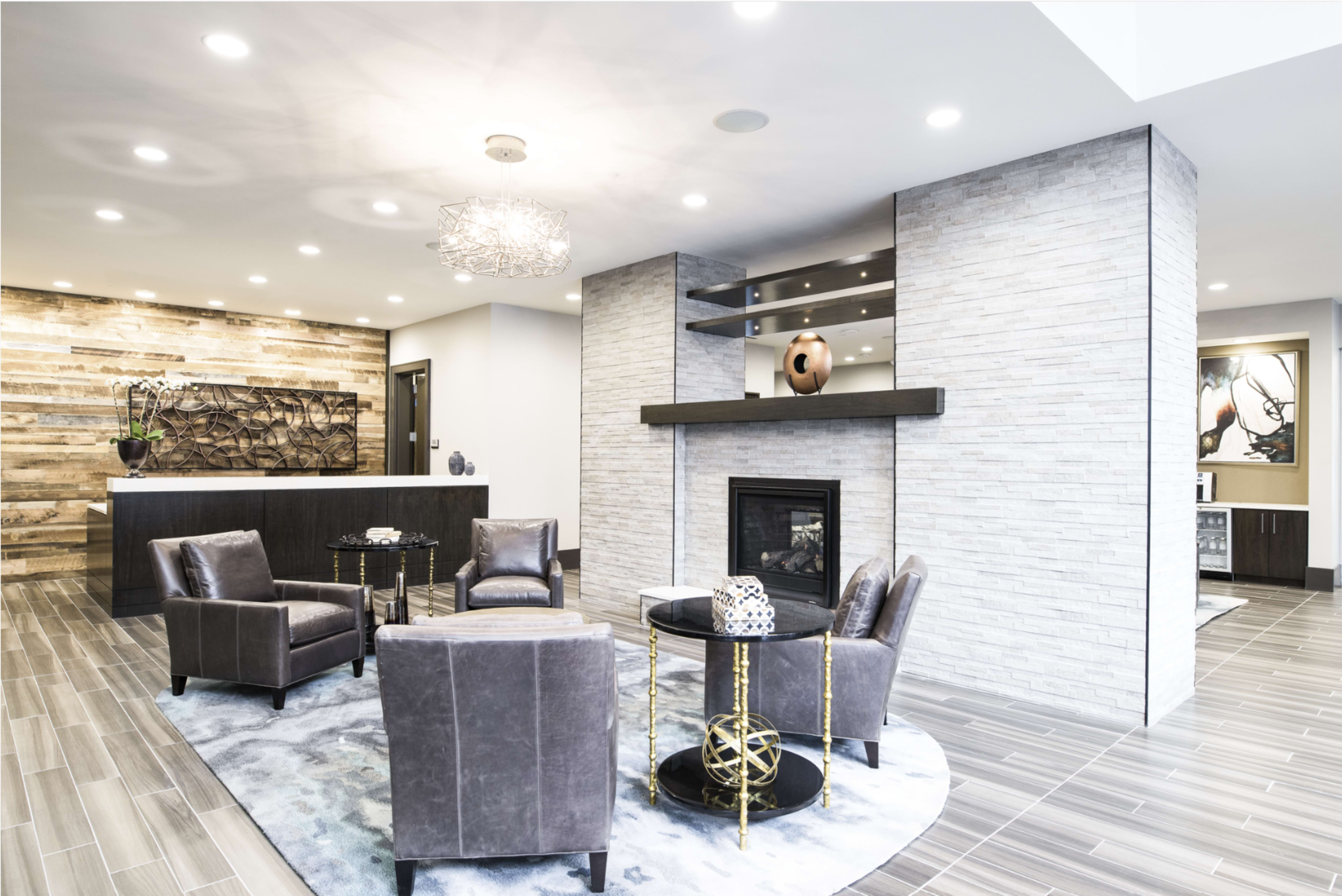 The image shows a modern lobby with a fireplace, seating area with four chairs, and a reception desk featuring a wooden wall behind it.