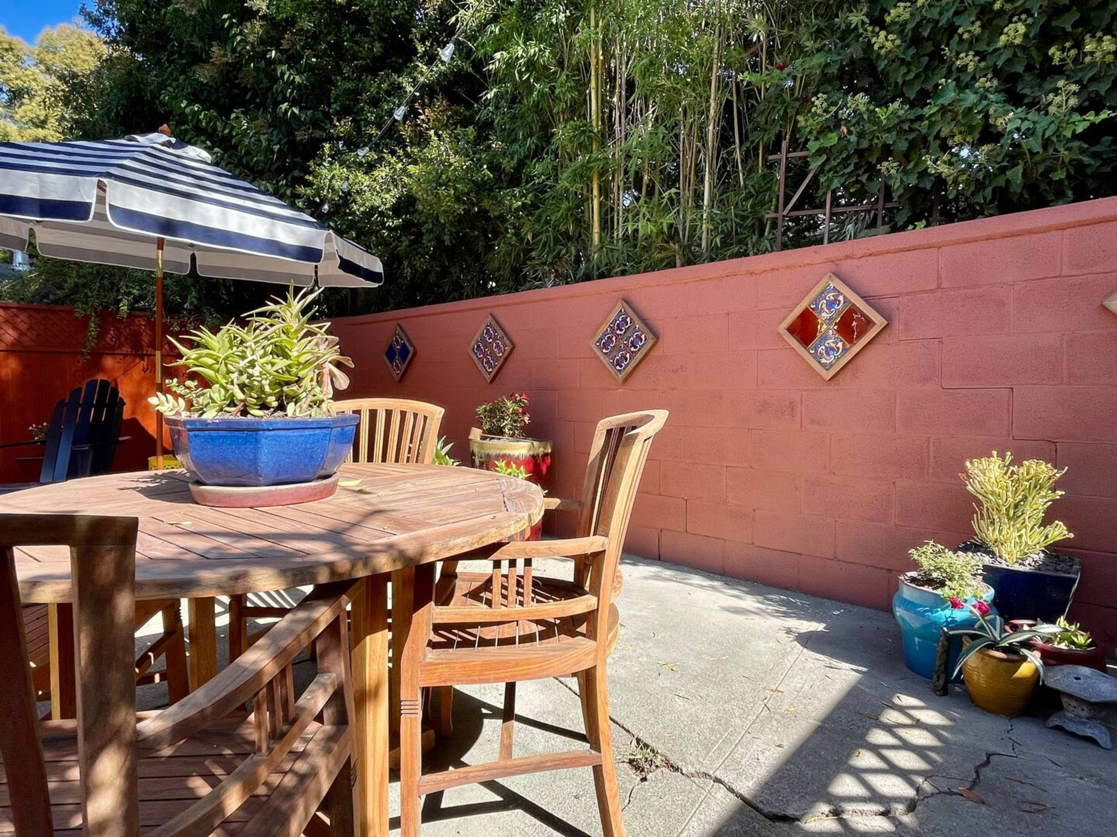 A wooden table and chairs are arranged on a patio beside a pink wall adorned with decorative tiles and potted plants.