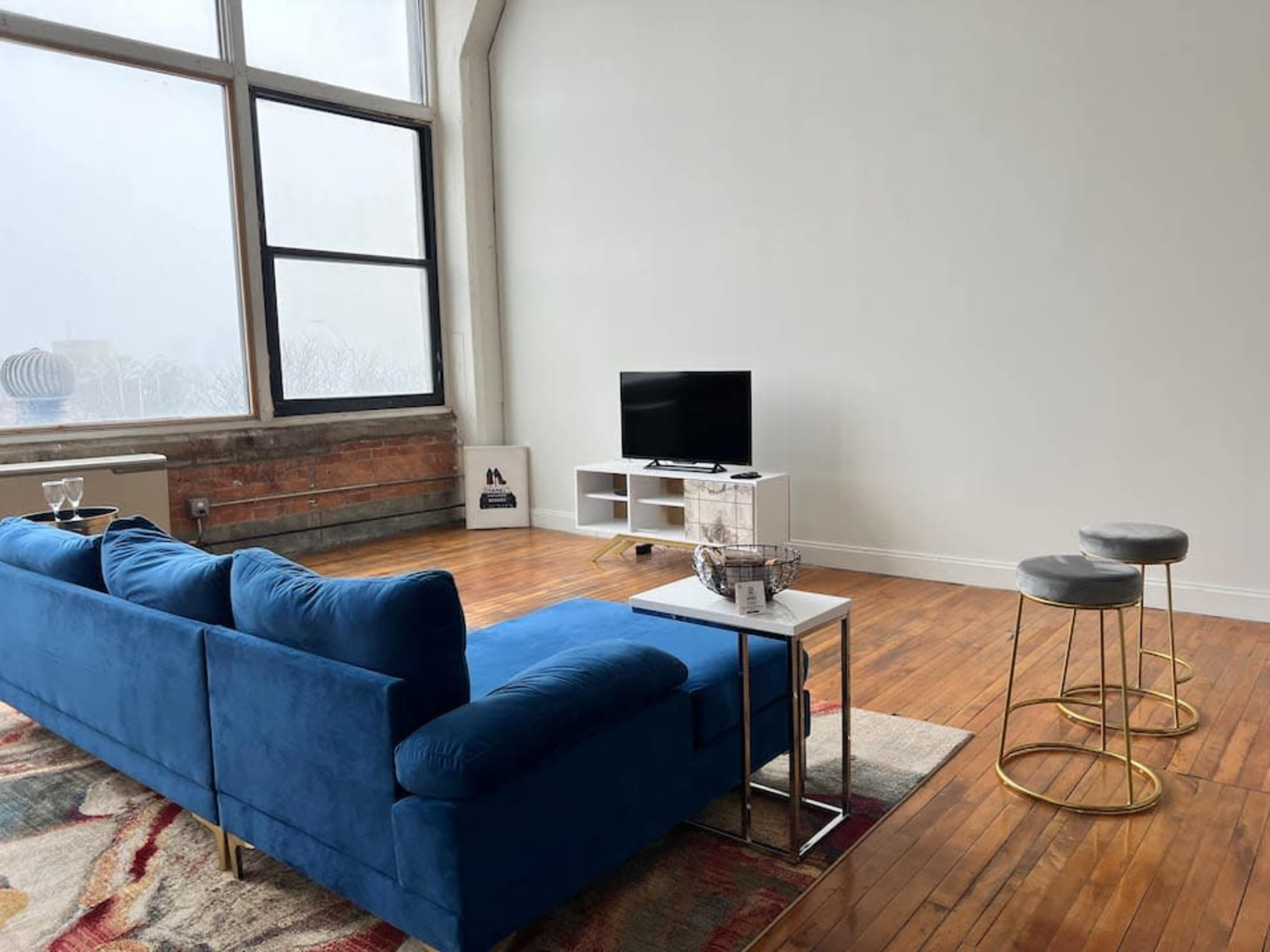 A blue sofa faces a television on a white stand in a spacious room with large windows and wooden flooring.