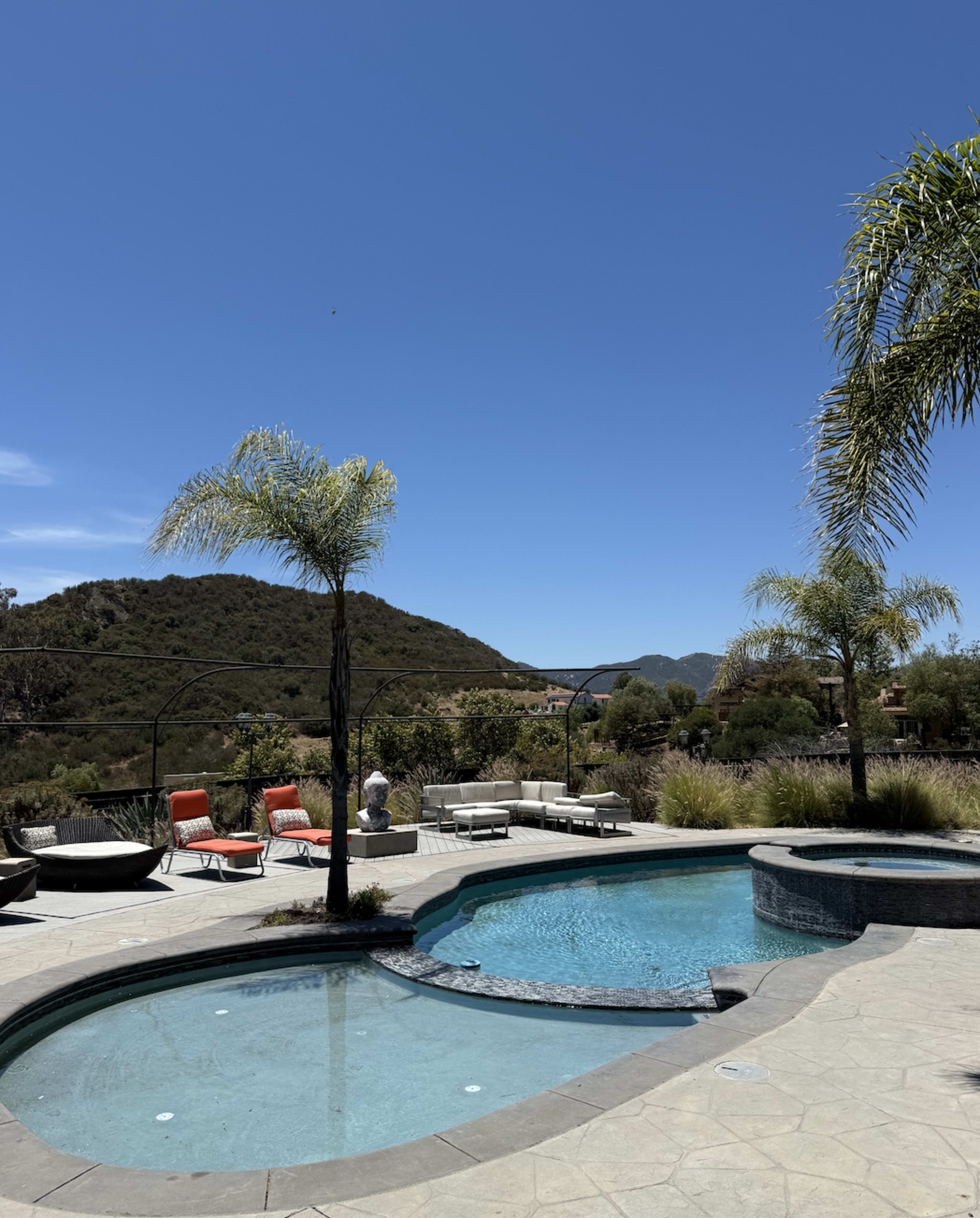 A swimming pool with a curved edge is surrounded by lounge chairs and palm trees, set against a backdrop of rolling hills and a clear blue sky.