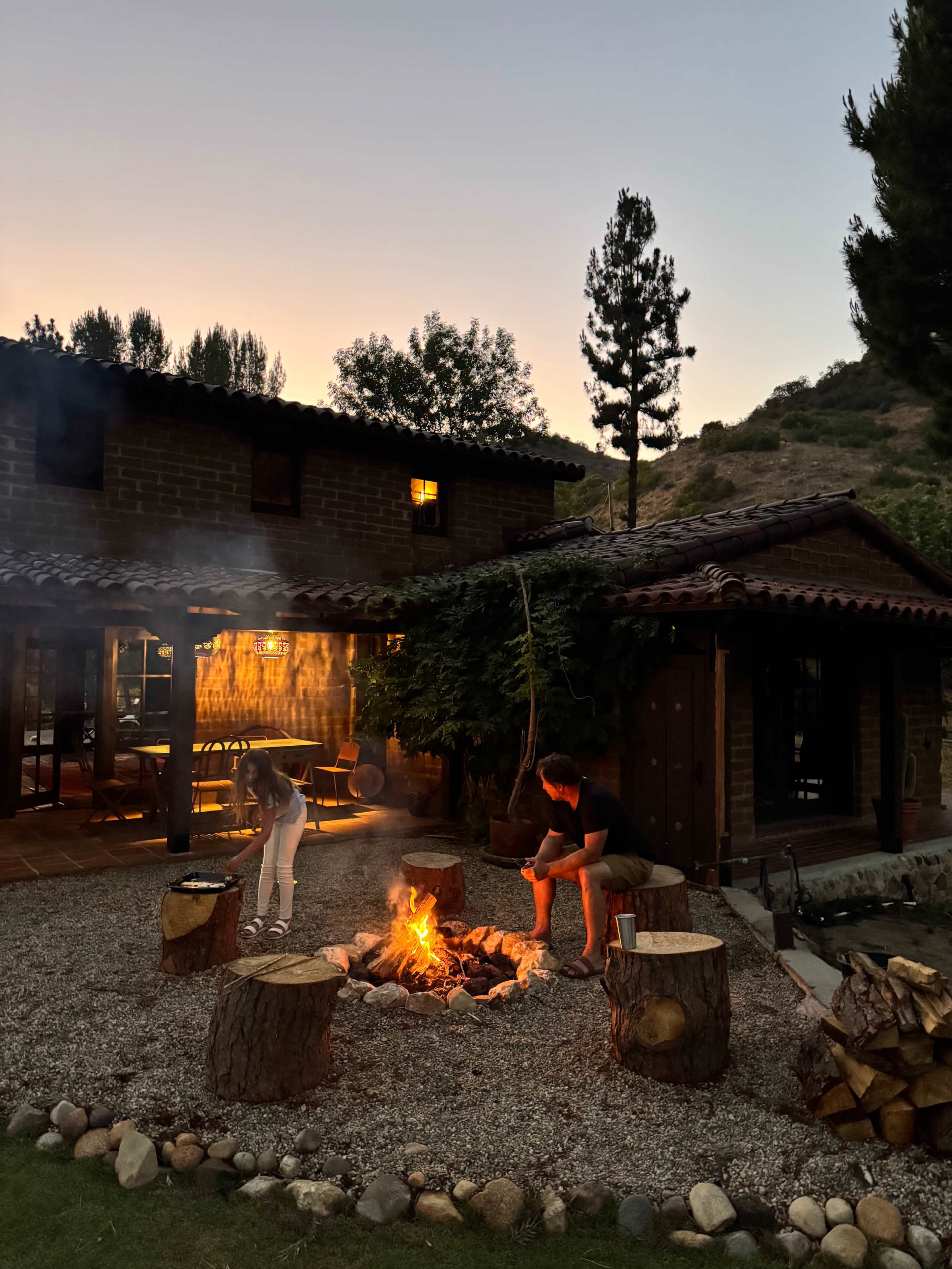A person sits by a campfire in front of a rustic cabin as twilight descends over the surrounding landscape.