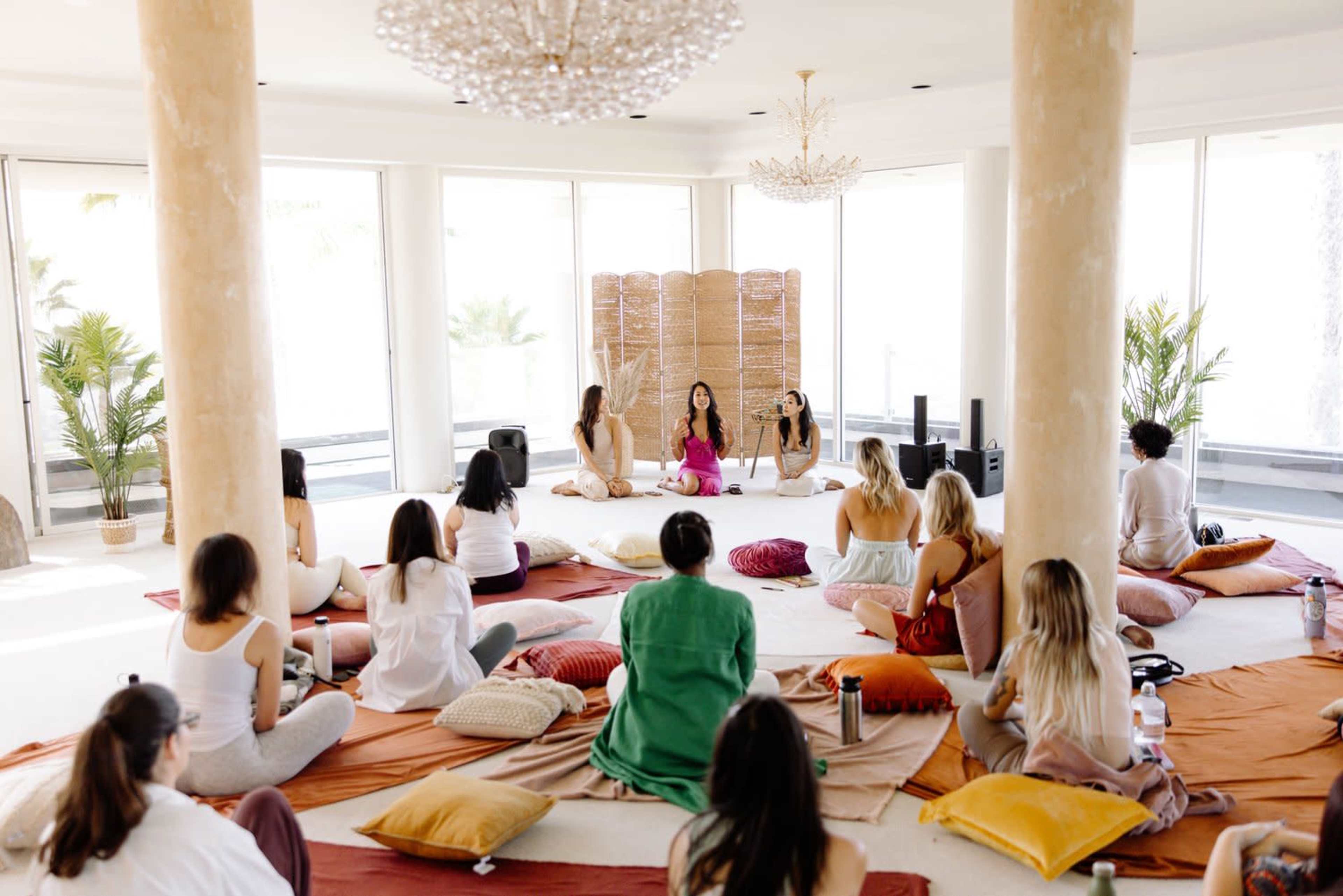 A group of people sits on floor cushions in a bright, open space while a few individuals lead a discussion in front of a bamboo screen.