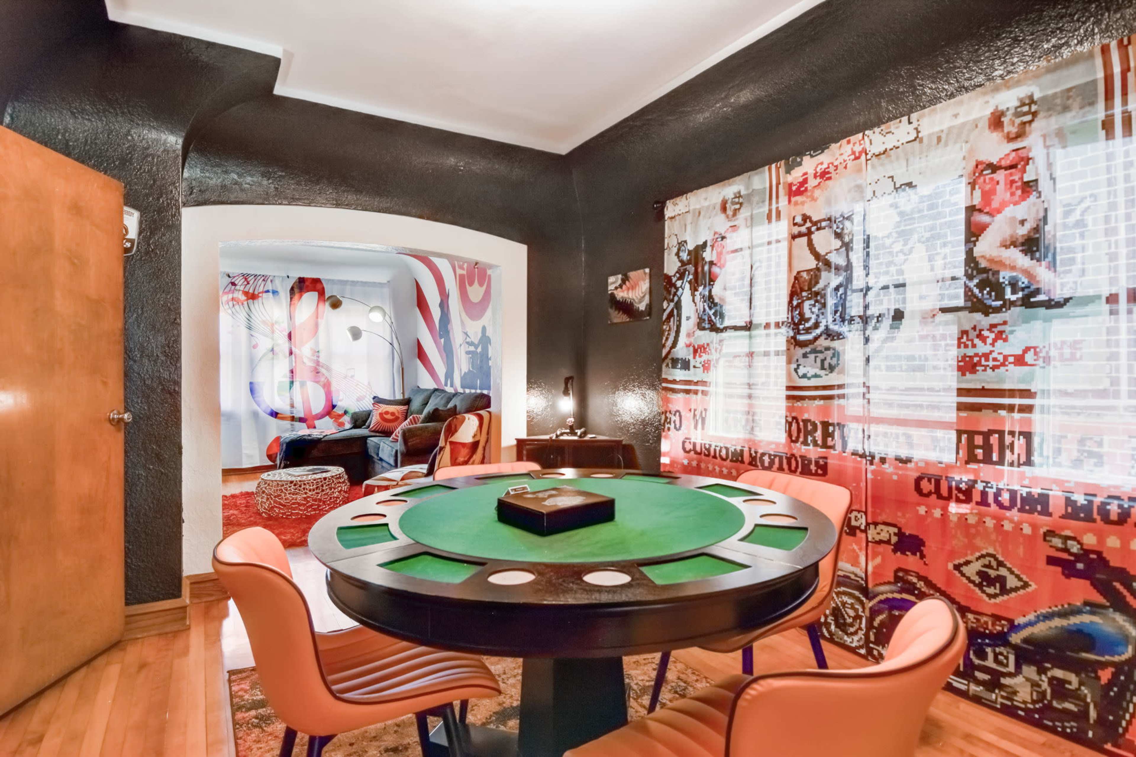 A brightly decorated game room featuring a circular green poker table surrounded by orange chairs, with colorful artwork on the walls and patterned curtains.