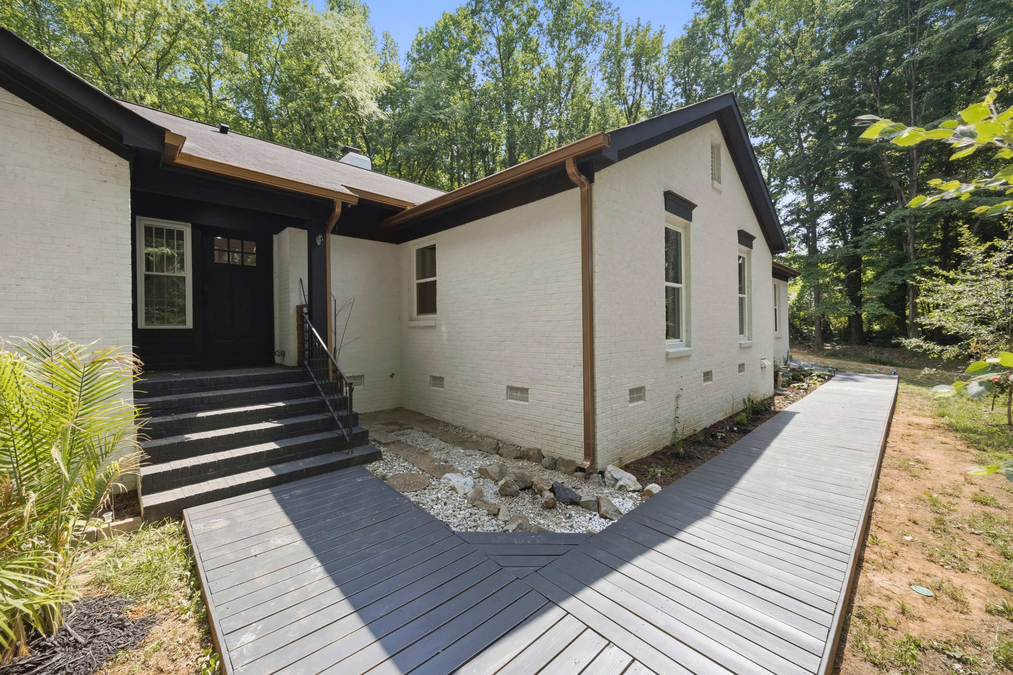 The image shows a modern white house with a dark front door and a wooden walkway leading to its side, surrounded by greenery.